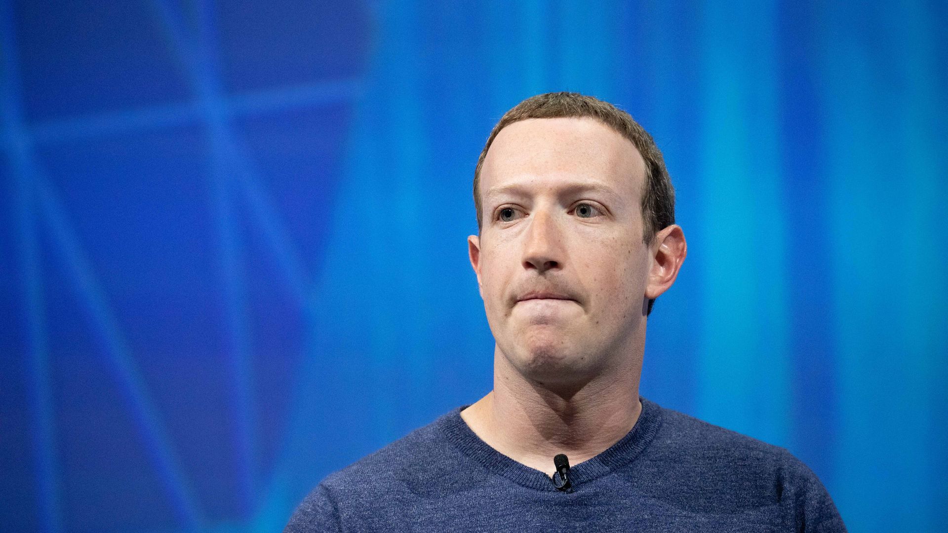 Mark Zuckerberg wearing a blue shirt before a blue background bites his lips frowning.