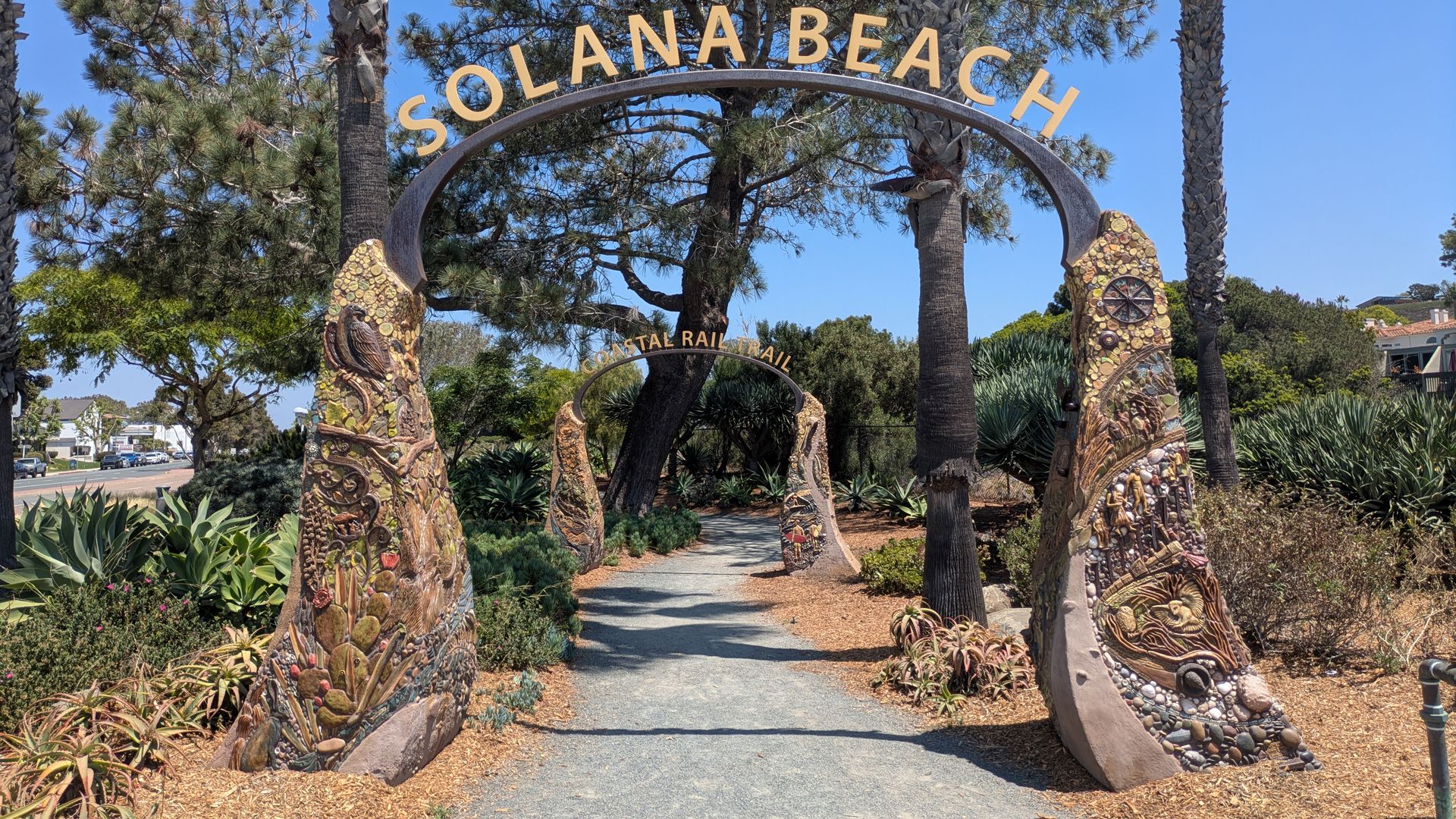 Entrance arch sign reading SOLANA BEACH over a curved metal frame, flanked by tall palm trees; gravel path and desert landscaping under a blue sky.