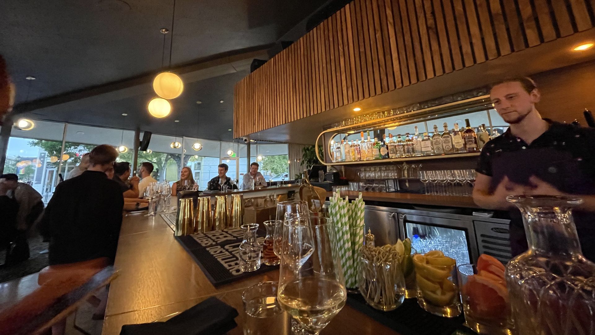 A man stands behind a bar with numerous bottles behind him, and glassware and other materials in front of him. 