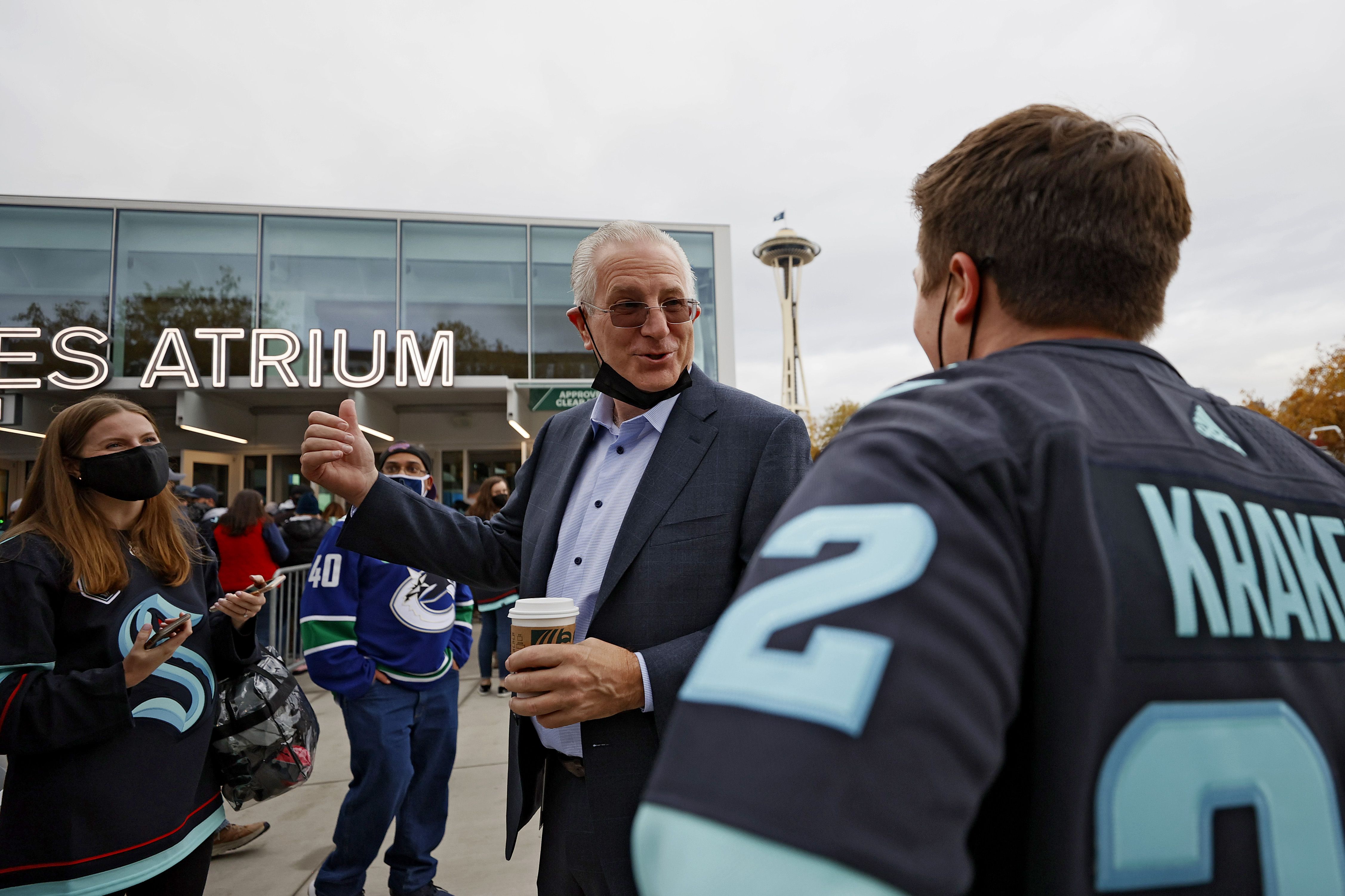 A man holding a coffee cup talks to people in jerseys outside. The Space Needle is in the background.