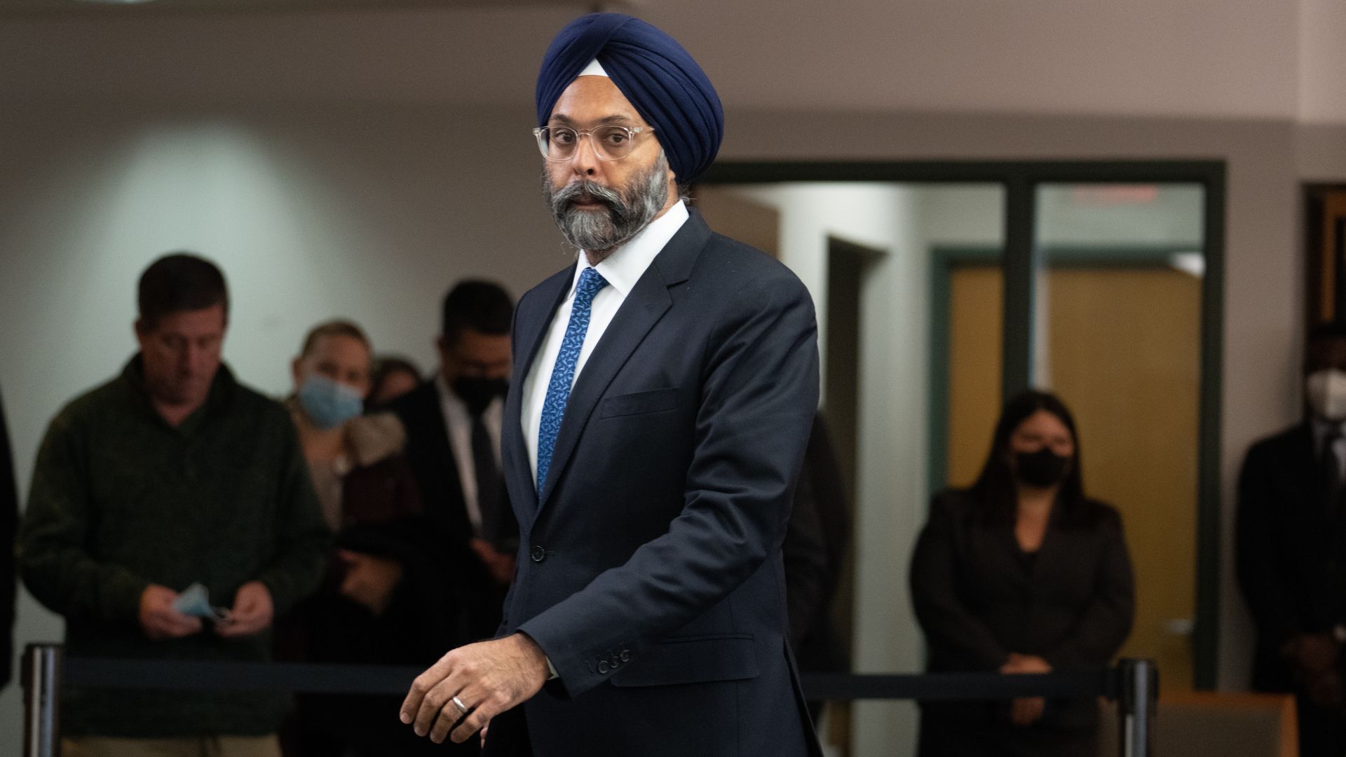 A man in a blue business suit and matching turban walking into a room with a number of people in shadows in the background looking on. 