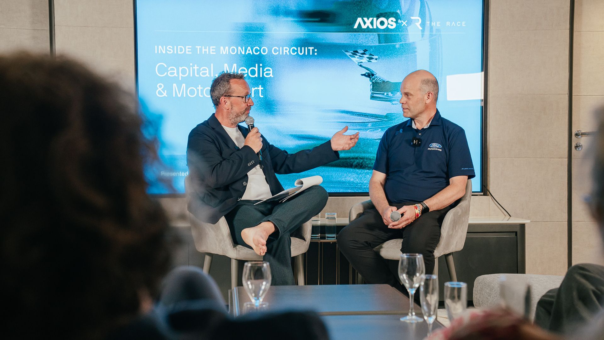 Two men sit on a panel stage; left speaker gestures with a mic, right listens in a navy polo. A blue screen behind shows "Inside the Monaco Circuit: Capital, Media & Motorsport".