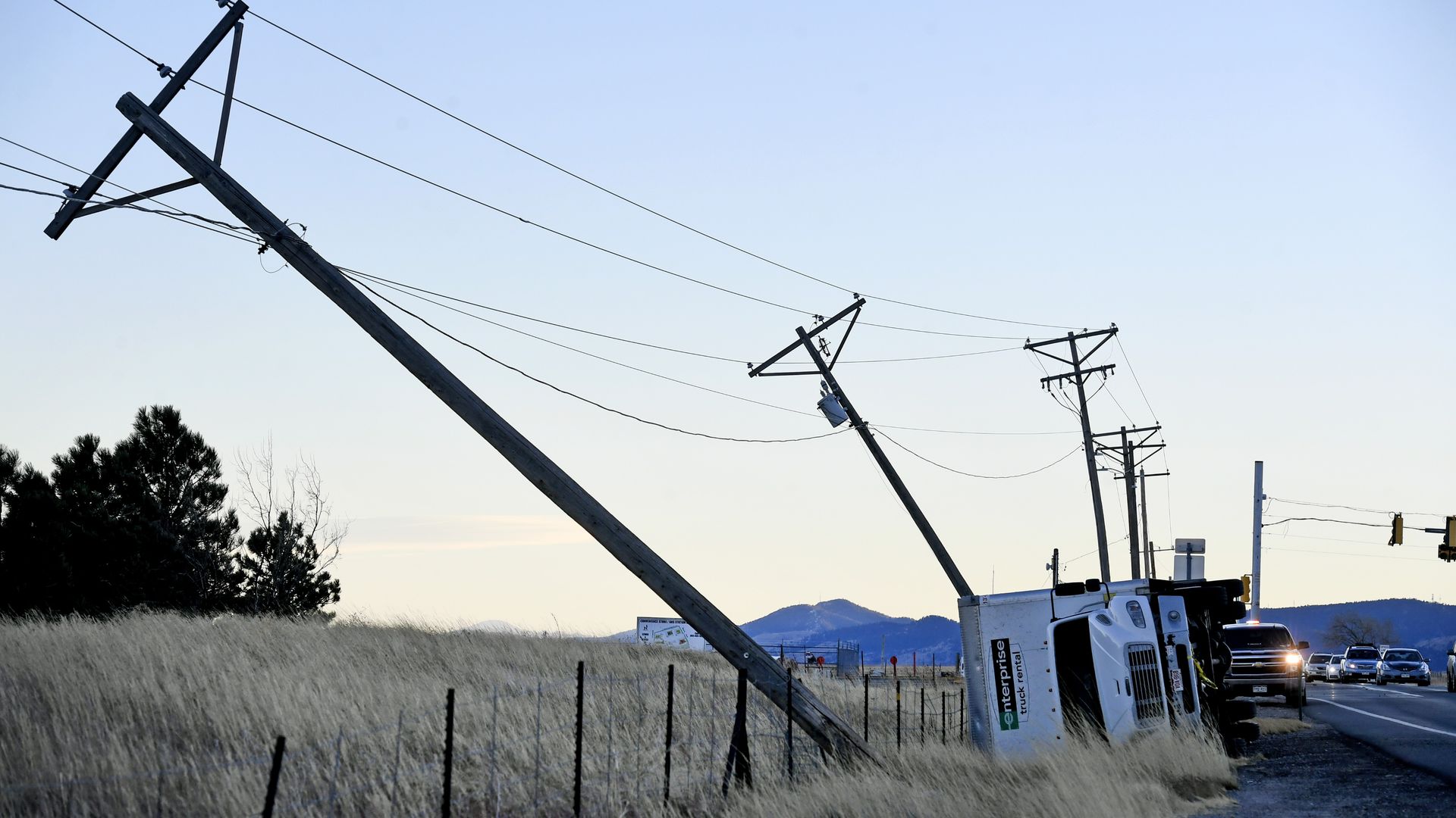 White Enterprise truck on its side beside a road with leaning utility poles and power lines in a dry grassy area during early evening with hills in the background.