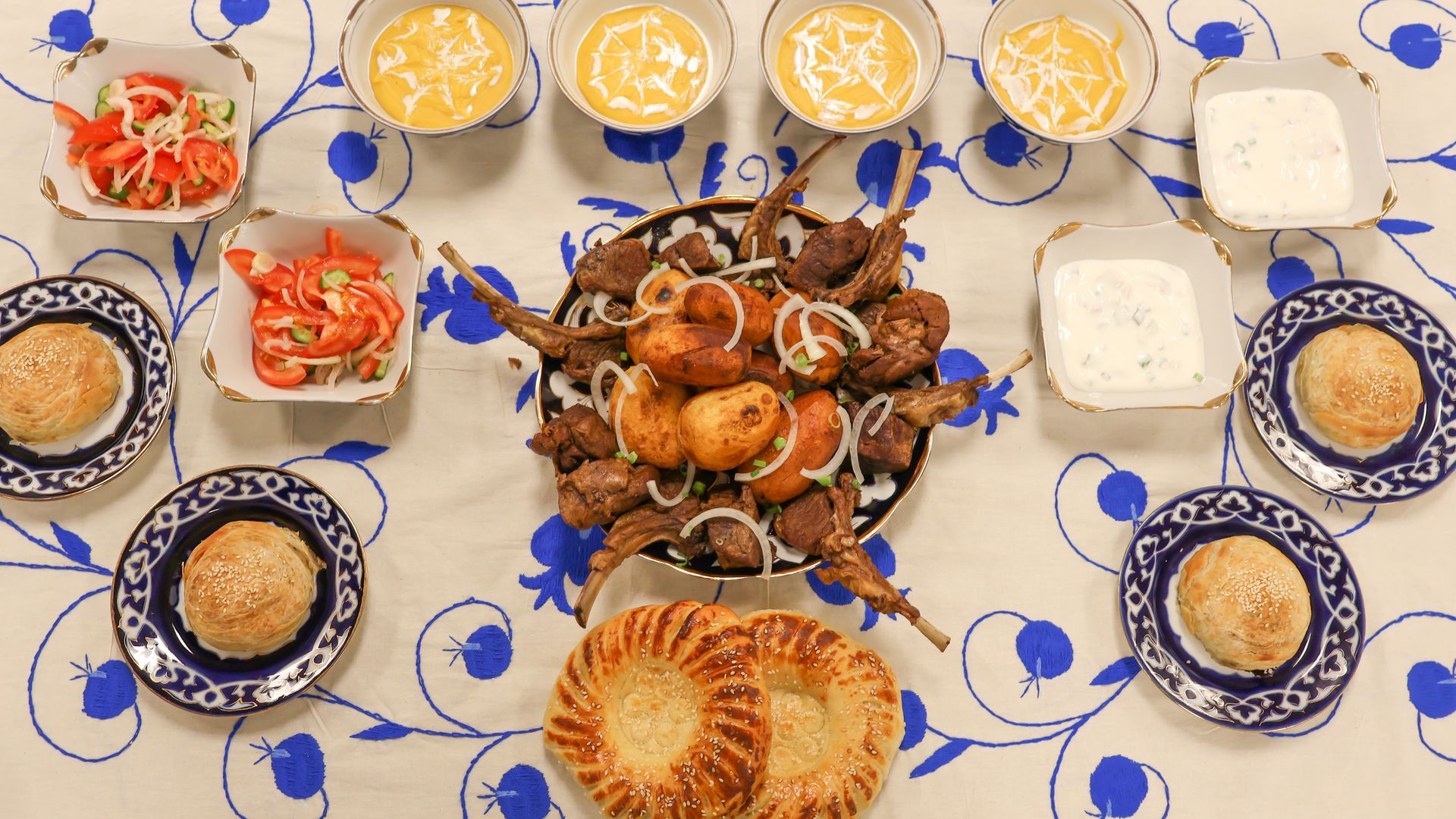 A table set with a central dish of grilled meat and potatoes garnished with onions, surrounded by bowls of yellow soup, salad with tomatoes and onions, yogurt sauce, and round bread on blue-patterned plates.