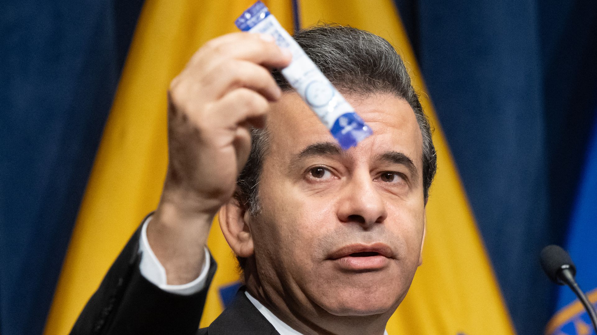 Man in suit and blue tie holding up a blue and white packet, with yellow and blue flags in the background, speaking at a microphone.
