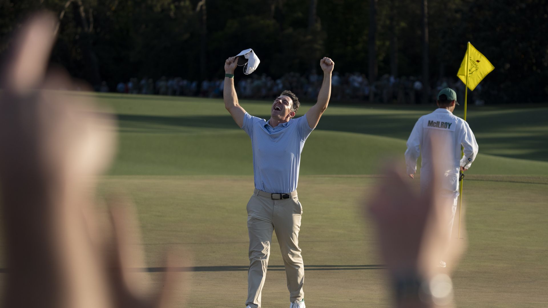 A golfer wearing khaki pants and a light blue shirt holds a white hat in his hands and his arms over his head in victory on a putting green. A caddy in white coveralls with a patch that says "McIlroy" and wearing a green hat is holding a yellow pin flag.