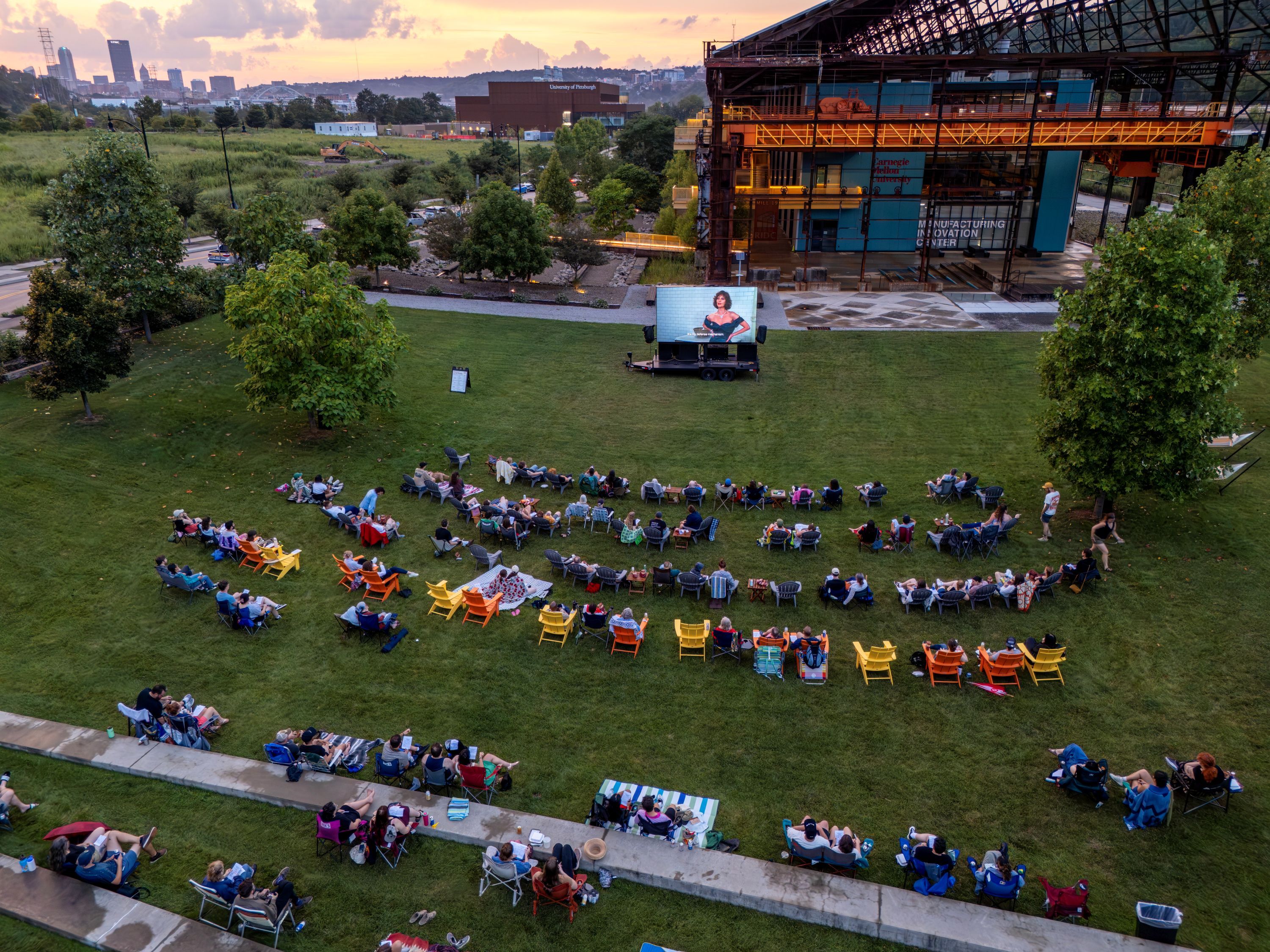 An aerial view of an outdoor movie night on a grassy hill at Carnegie Mellon University. Screen shows a woman in red; rows of colorful chairs face it as sunset lights the city skyline.