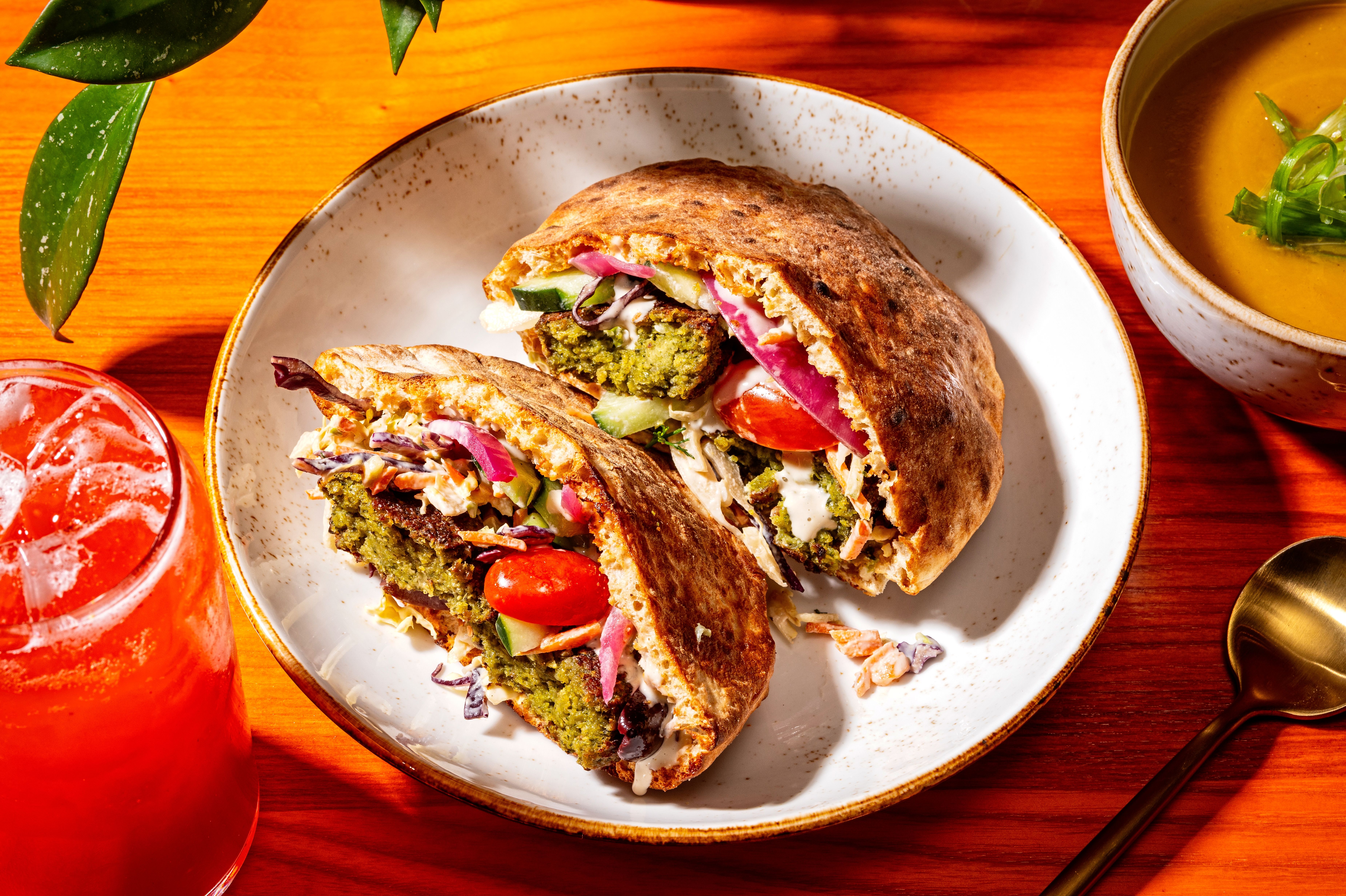 A falafel sandwich on a bright table with a red drink and green plant