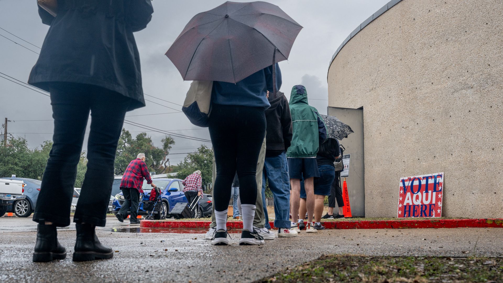 People stand in a line in the rain with umbrellas beside a beige, rough-textured wall; a VOTE HERE sign is visible to the right, with wet pavement and parked cars in the background.