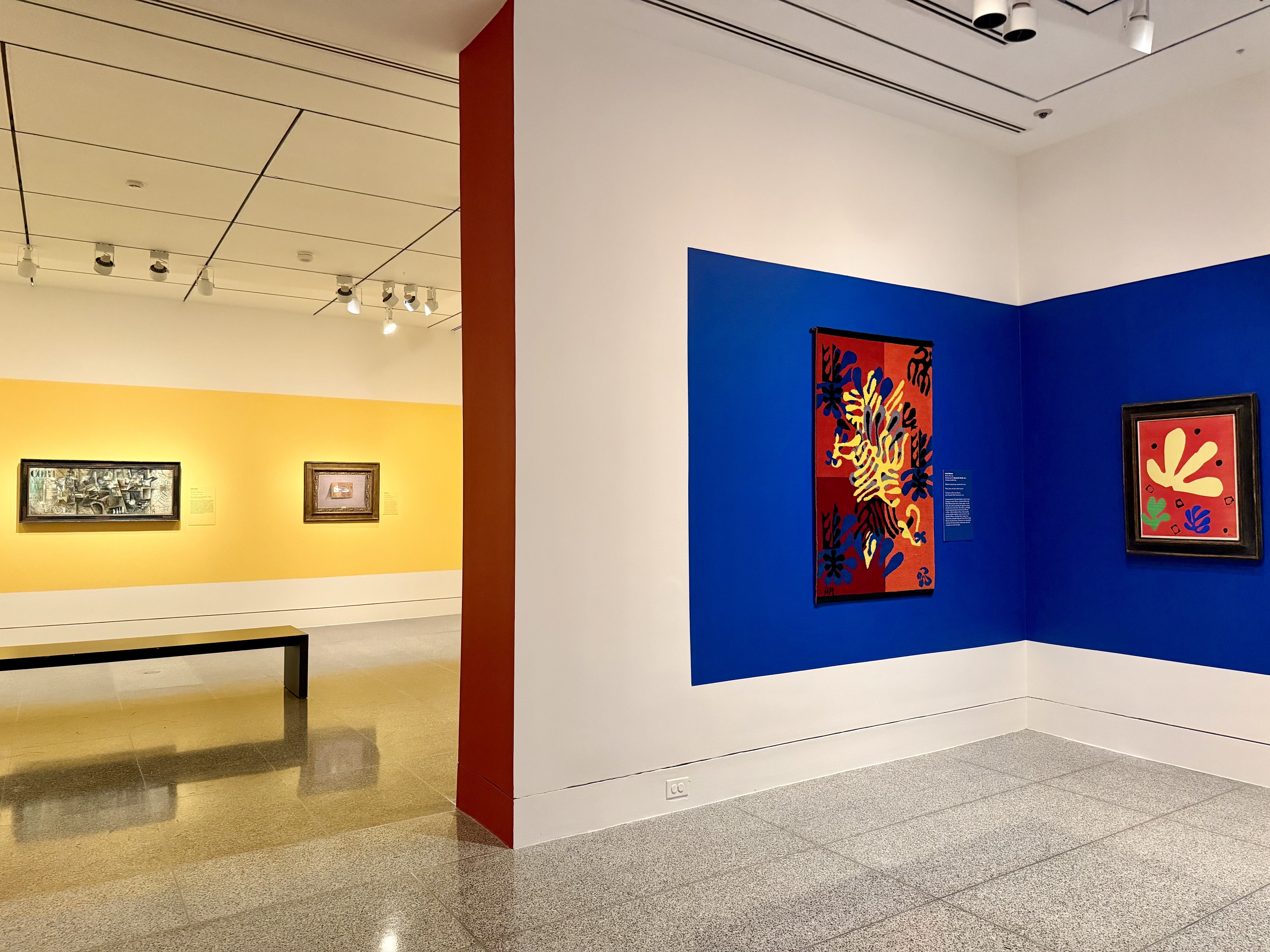 Interior of a modern art gallery: left yellow wall with two small framed paintings, right blue wall with large colorful works; red central column, white ceiling, track lighting, glossy floor.