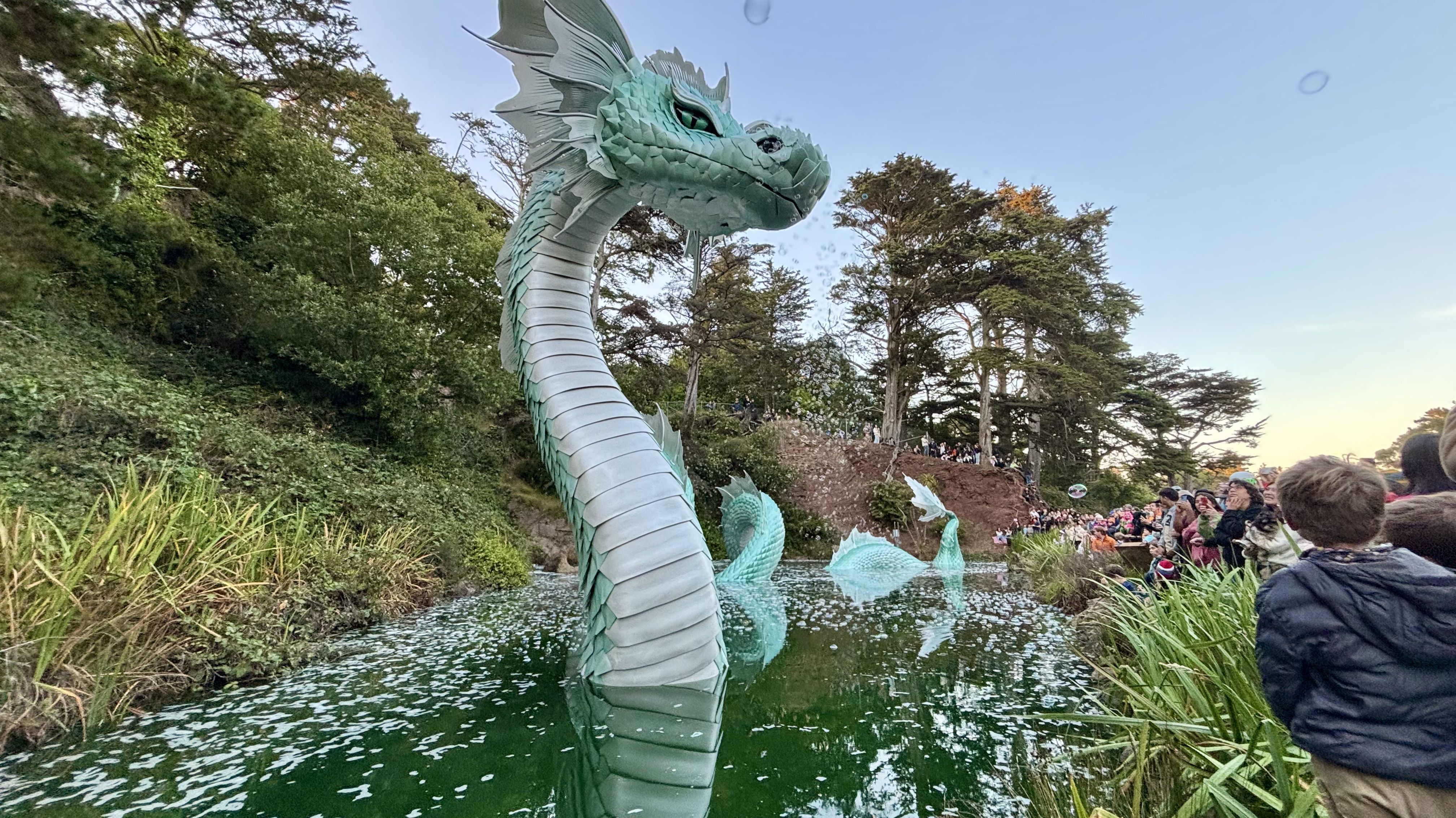 Large green serpent sculpture rising from a pond surrounded by trees and crowd of people watching during clear daytime.