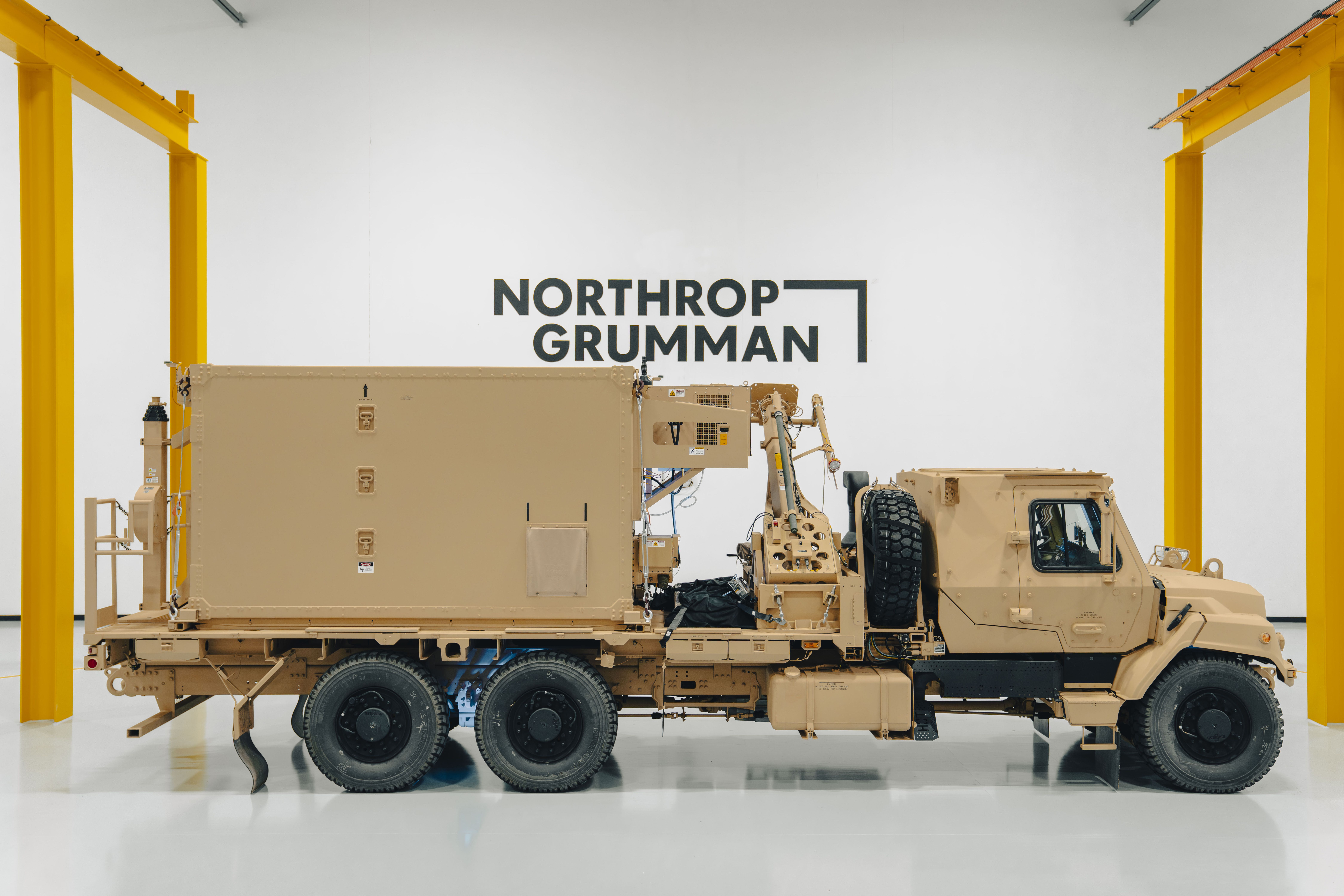 Side view of a tan military truck with six large wheels parked inside a bright, clean facility with yellow structural beams and a "Northrop Grumman" logo on the white wall.