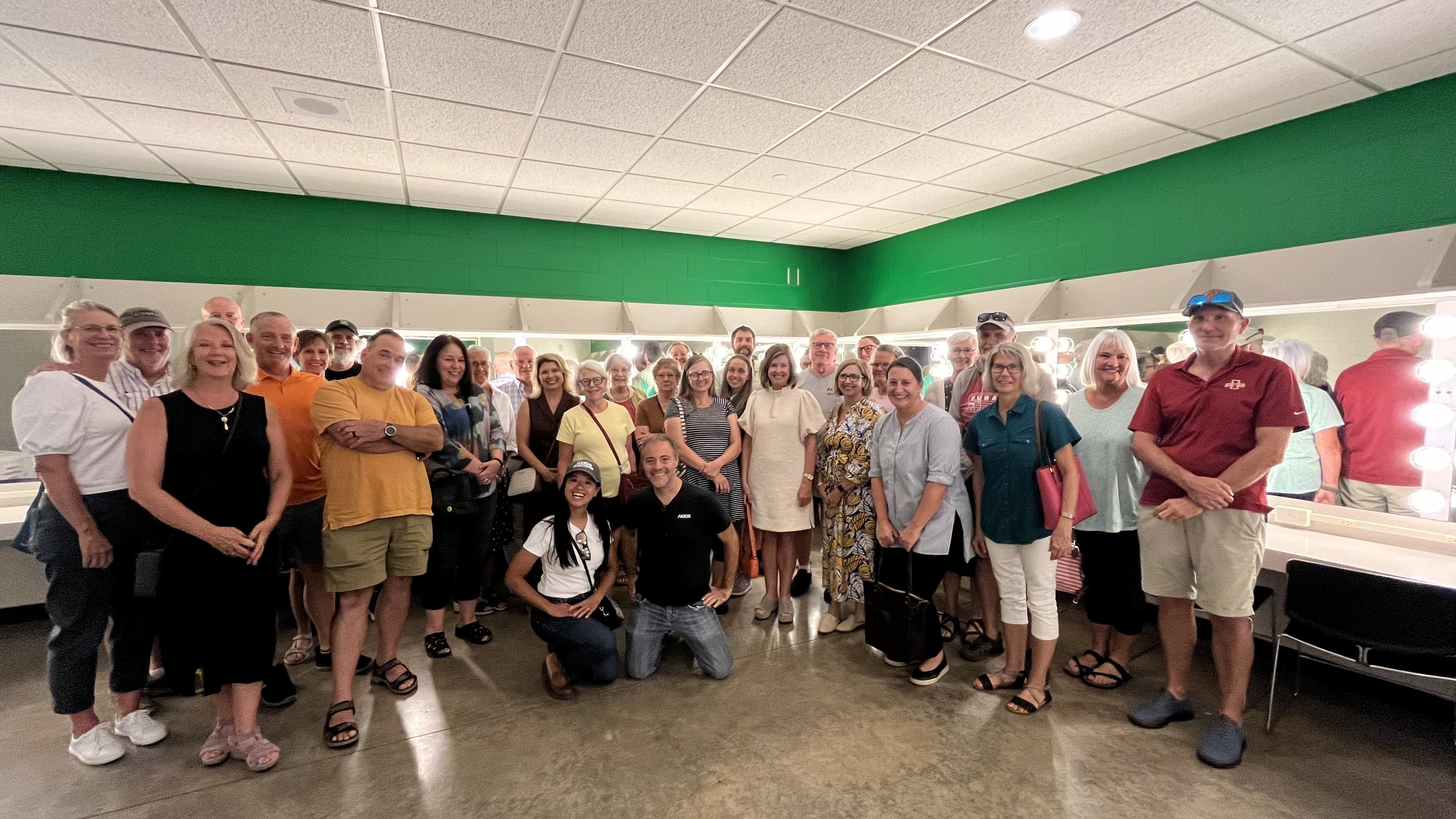 Large group of men and women posing in a green-walled room with vanity mirrors and lights.