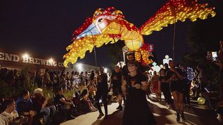 A woman carrying a large lantern sculpture leads a nighttime parade past a brewery and large crowd