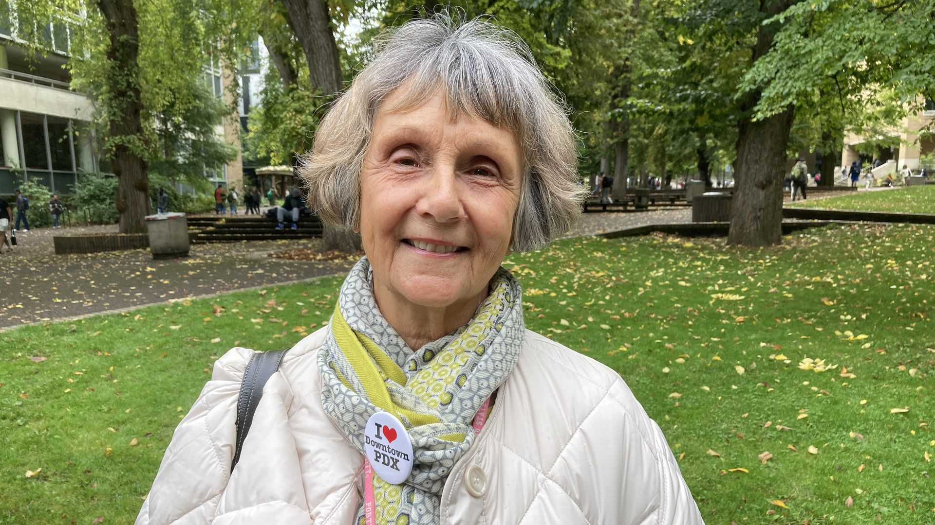 A smiling woman wearing a white coat and light green knitted scarf with a button that says I heart downtown PDX standing outside.