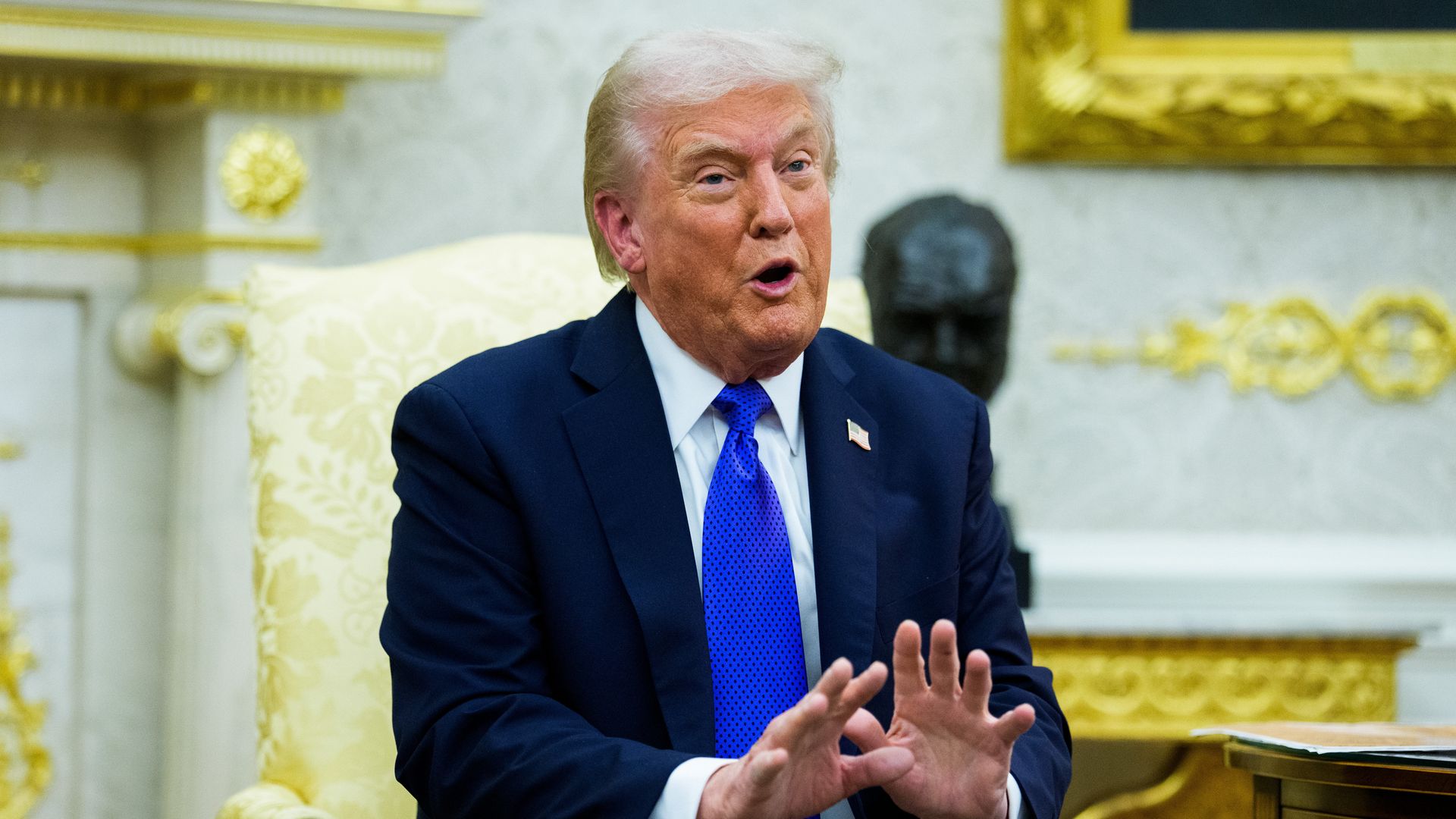 Donald Trump, wearing a blue suit and blue tie, sits in a gold chair in a white and gold room.
