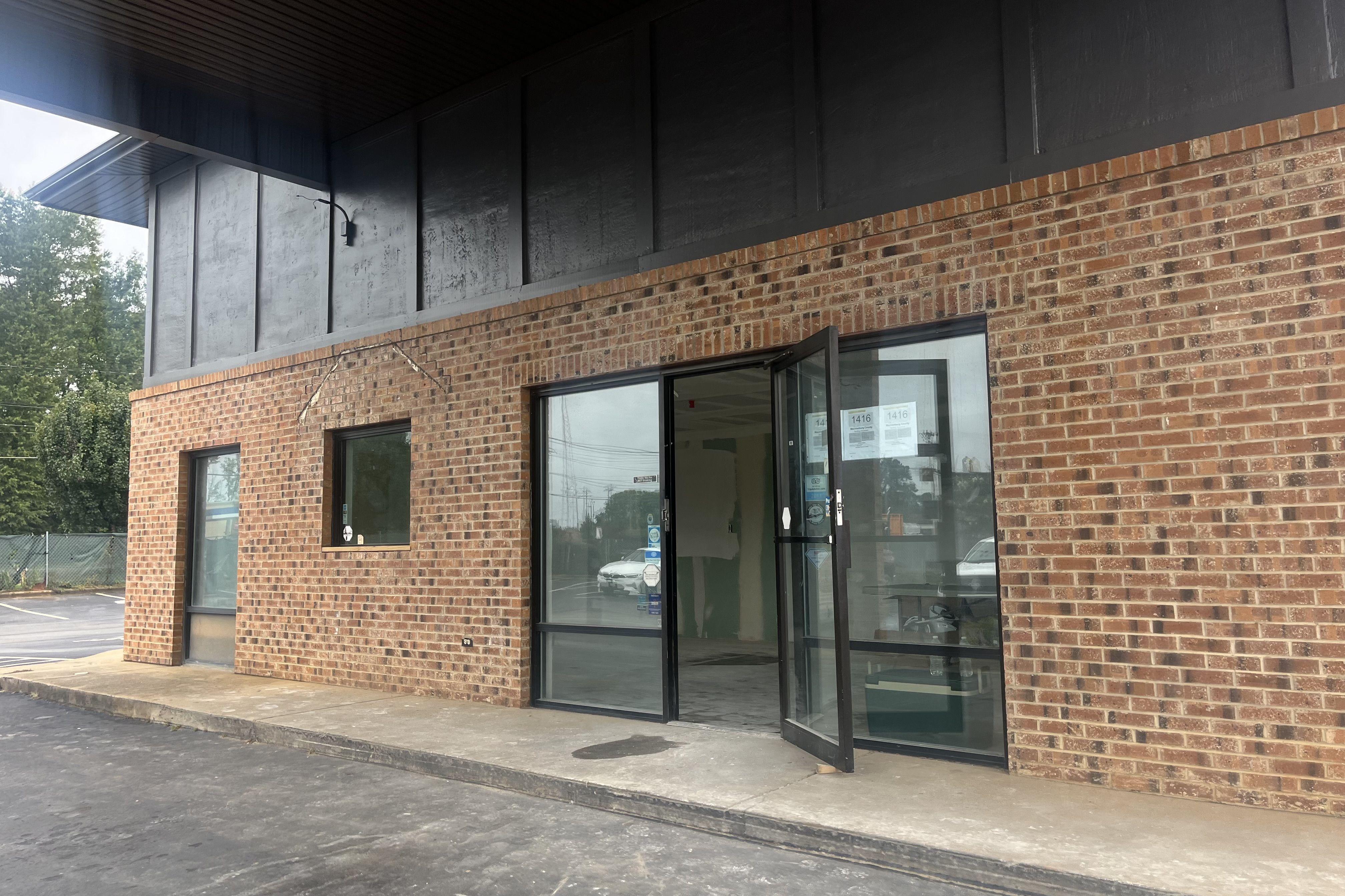 Exterior of a brick building with large glass doors and windows, one door partially open, under a black awning with a concrete walkway and parking lot in front.