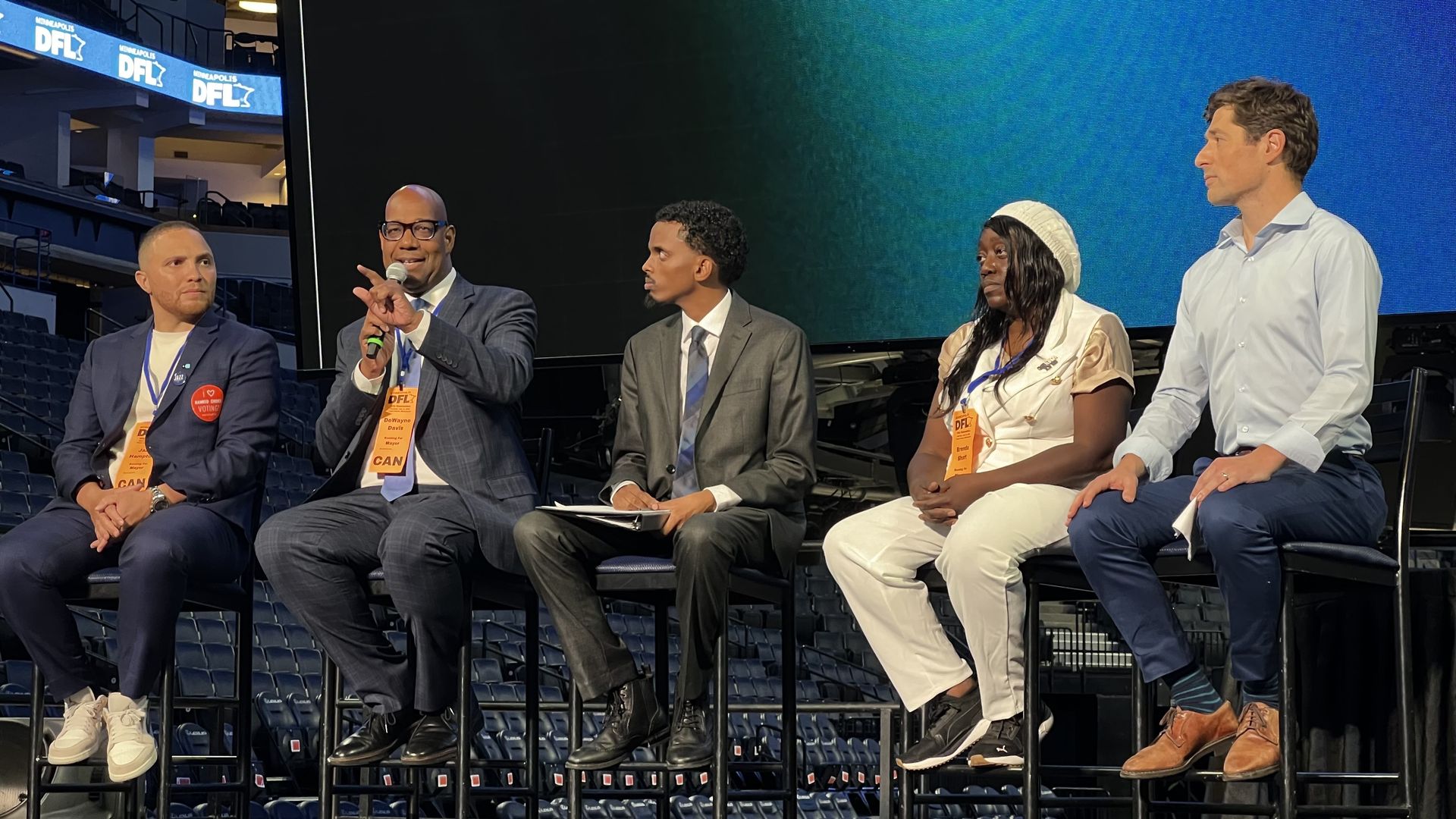 Five people sitting on a stage at a Minneapolis DFL event, four men and one woman, all wearing badges and engaged in discussion. One man holds a microphone and speaks.