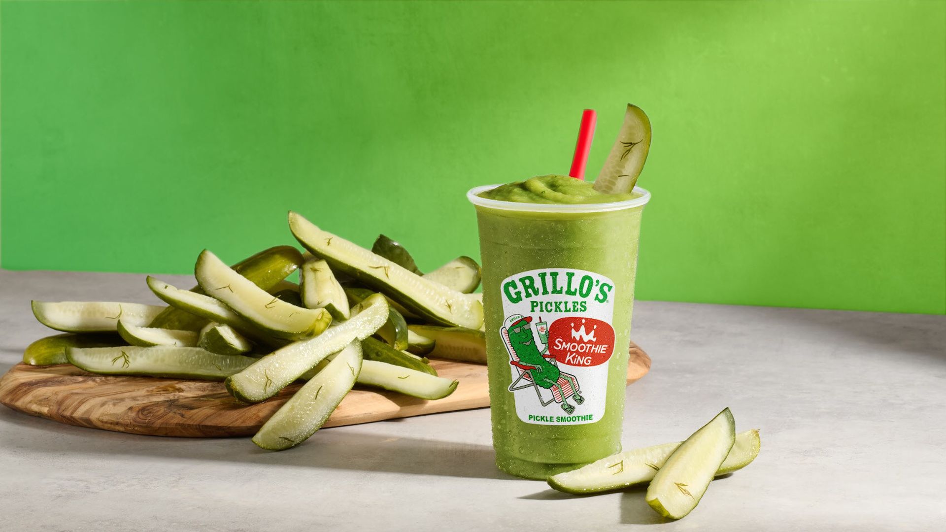 Bright green pickle smoothie in a cup with a red straw, on a gray surface, with a pile of pickle spears on a wooden board beside it; cup label reads Grillo's Pickles.