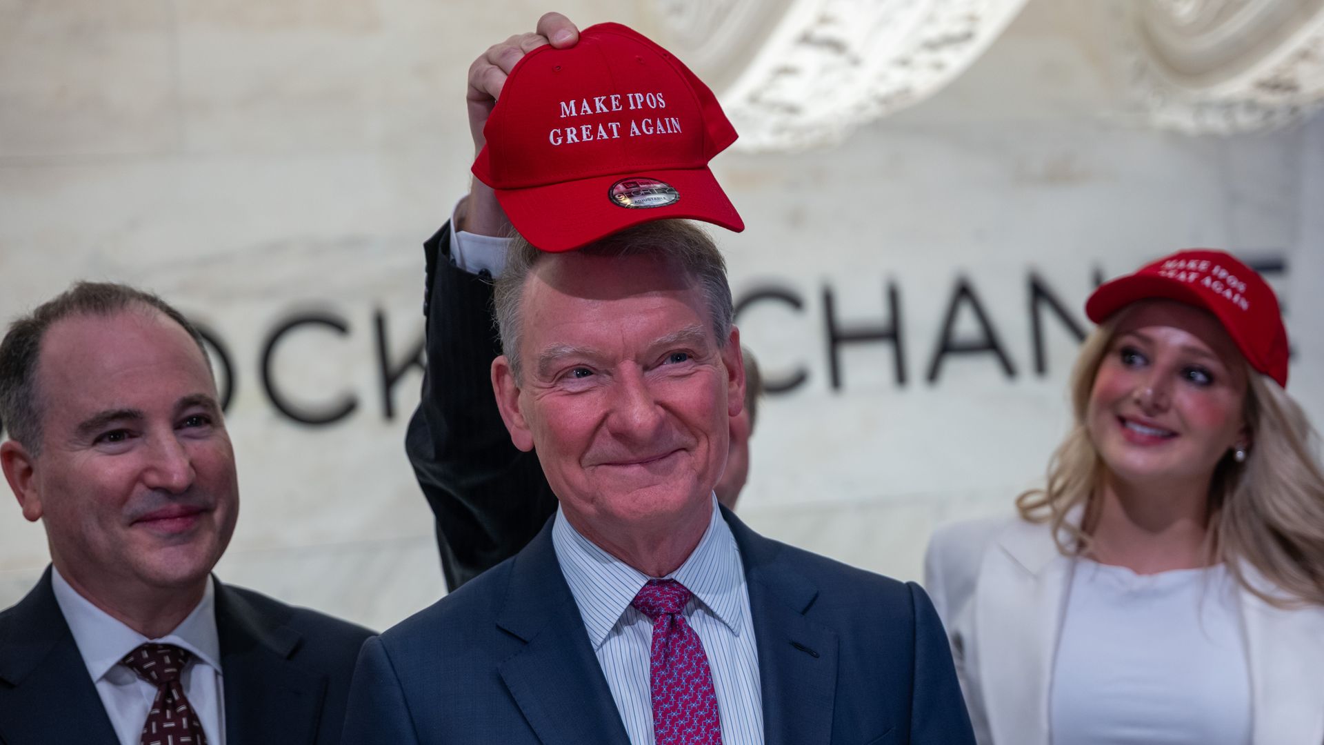 Smiling man in a dark suit raises a red cap reading "MAKE IPOS GREAT AGAIN" at a crowded event, with a blonde woman and another man nearby and a blurred backdrop.
