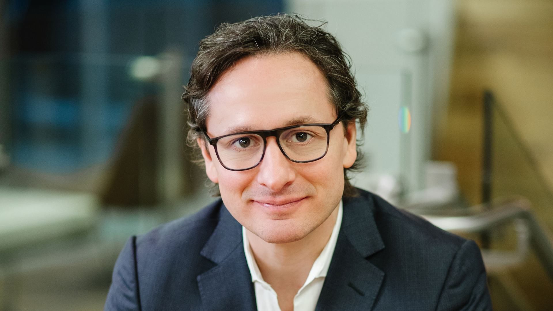 Portrait of a man with dark hair and glasses, wearing a navy blazer and white shirt, smiling softly at the camera in a modern, softly blurred office setting.