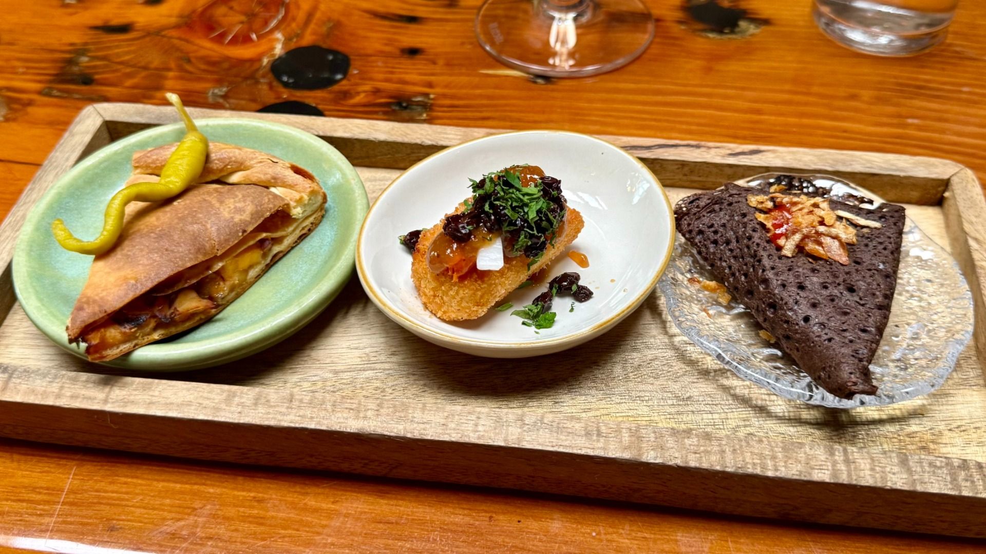 Three savory bites from MariPili’s tasting menu are arranged on a wooden tray, including a golden empanada, a croqueta with colorful toppings and a dark crepe garnished with crispy shallots.