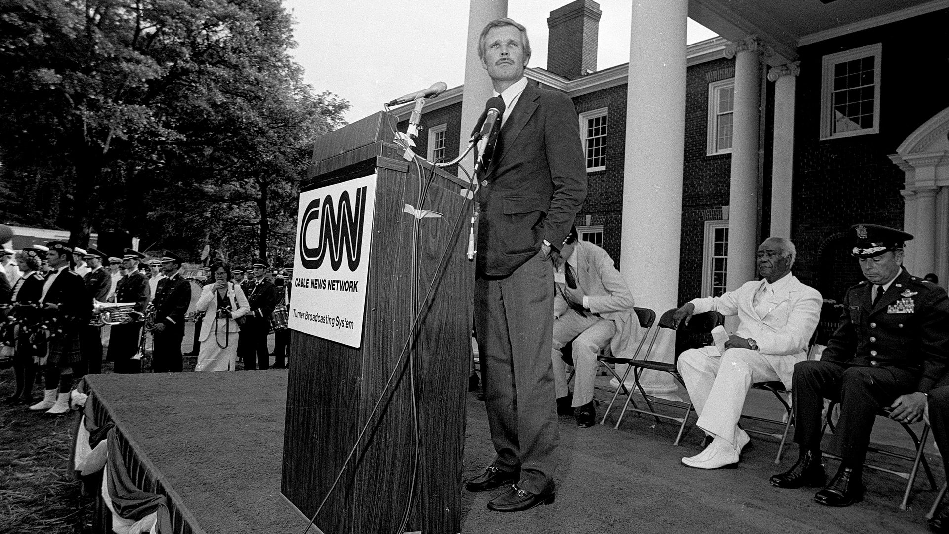 A man in a suit stands at a lectern on a podium in front of a columned building as a person wearing a white suit and a person in a military uniform sit in folding chairs behind him. A sign on the lectern says CNN.