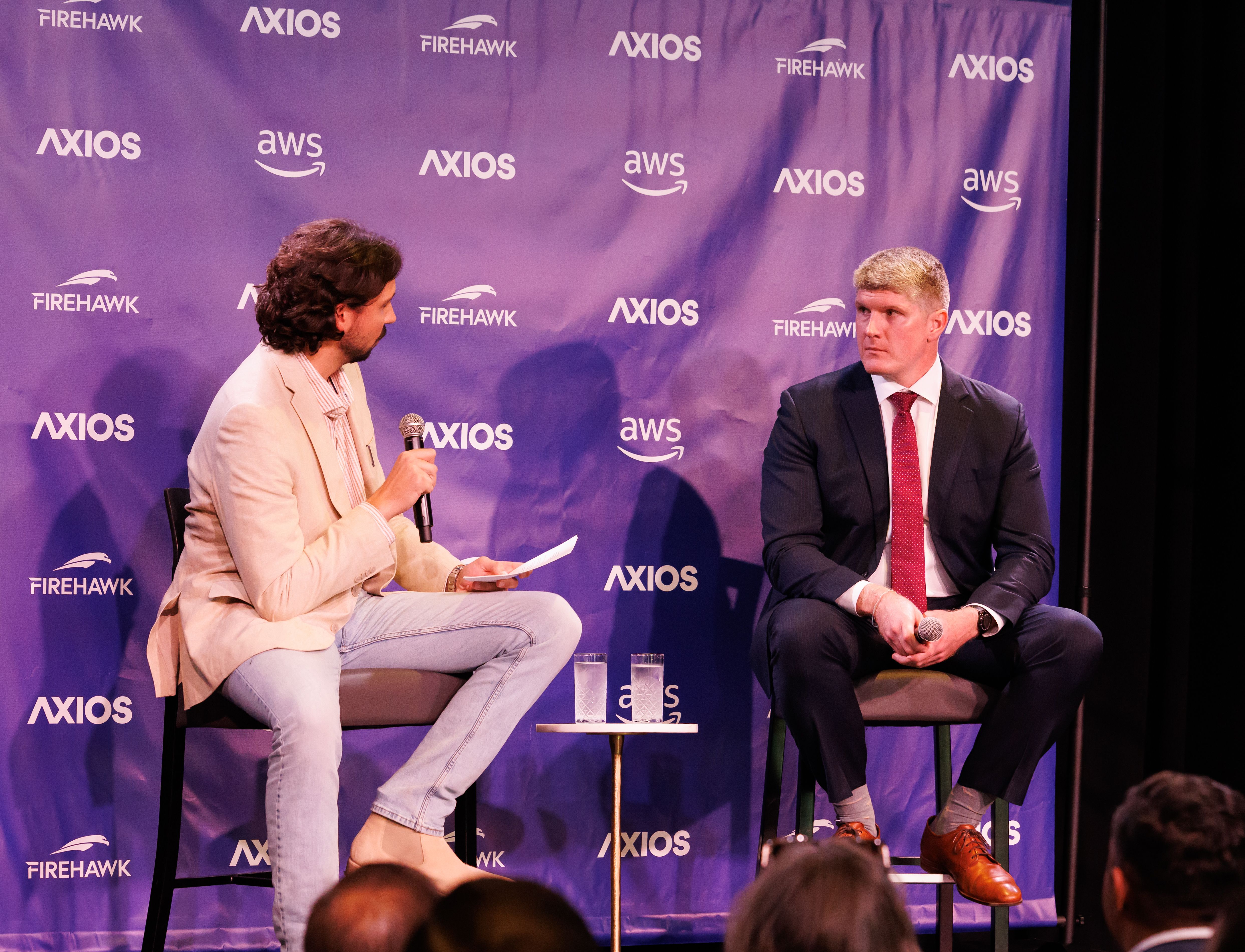 Two men sit on stage with microphones at an Axios event. One in a light beige blazer and jeans holds a paper, the other in a dark suit with a red tie listens. Blue backdrop with logos.