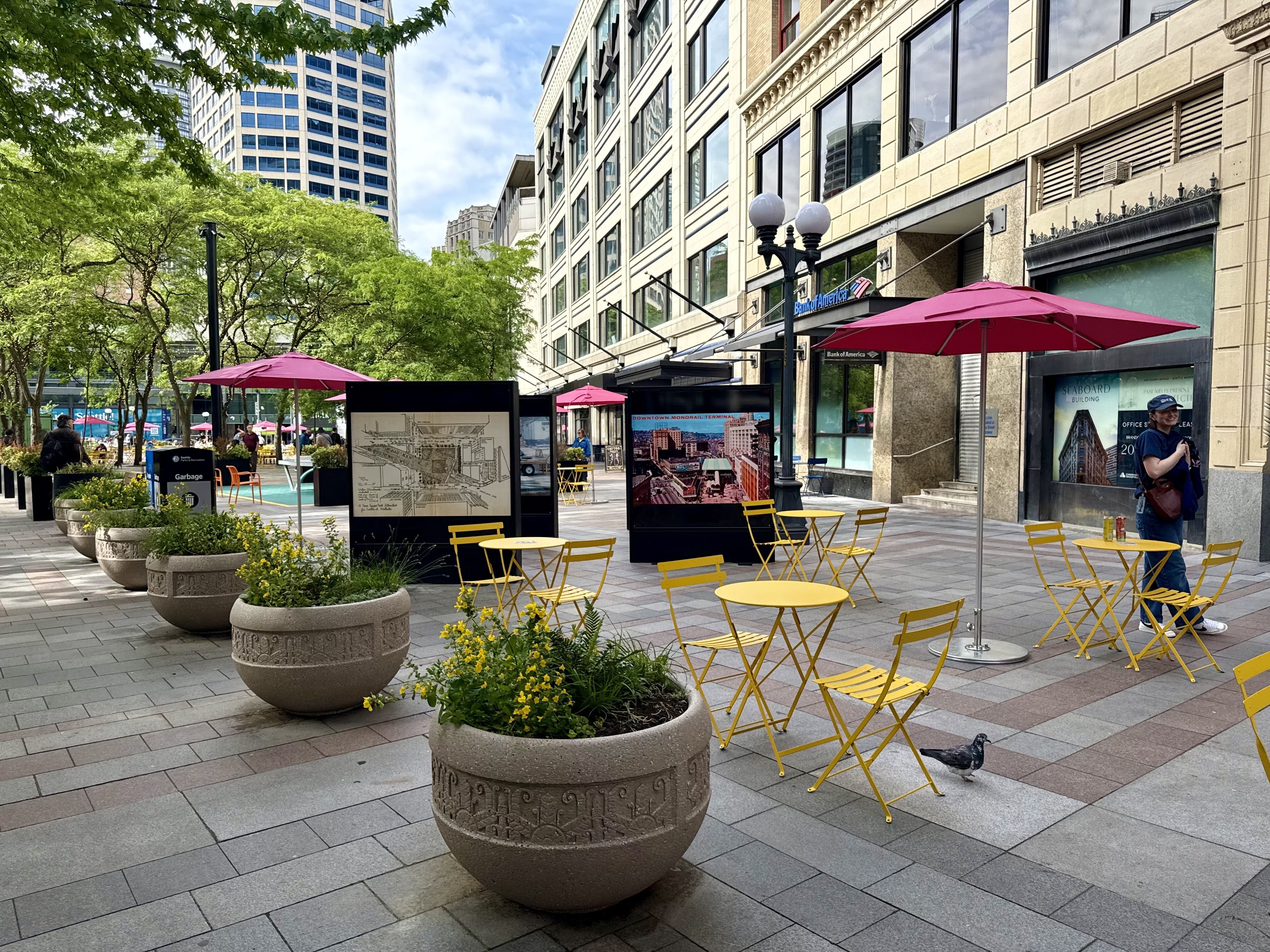 Urban plaza with bright yellow tables and chairs, pink umbrellas, and large planters. Green trees line the left; beige multi‑story building with many windows on the right, Bank of America sign, and a few people nearby.
