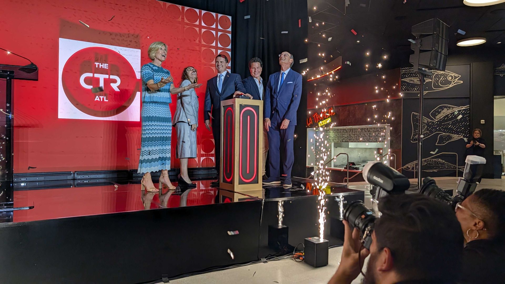 Group of formally dressed people on a stage with a red backdrop reading "The CTR ATL". Two women in blue dresses and three men in suits surround a lit podium as photographers film amid sparks.