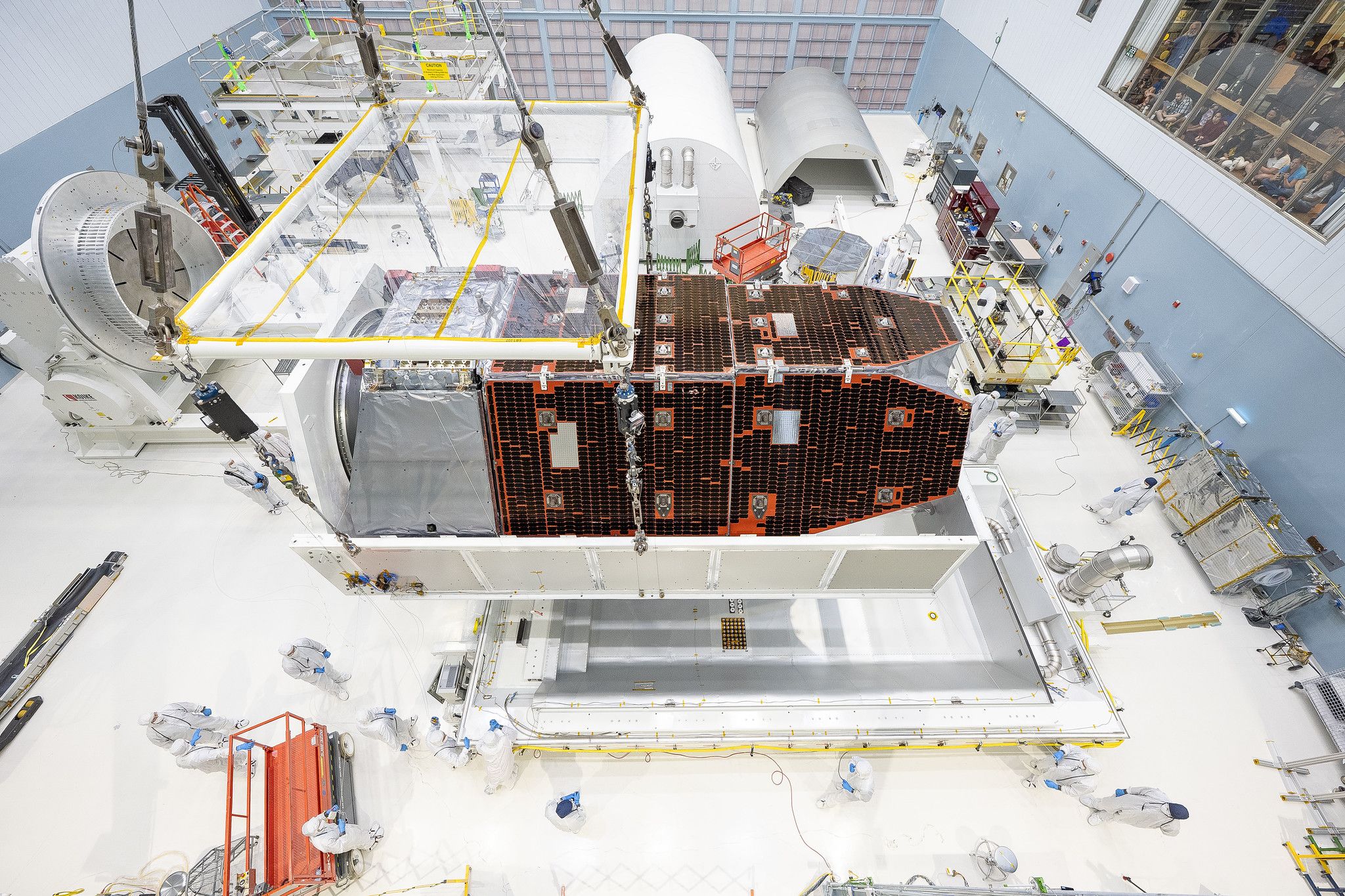 A wide cleanroom for space engineering shows a large satellite assembly: brown tiled solar arrays, white-suited technicians, orange scaffolding, yellow safety rails, and an observation deck with spectators.