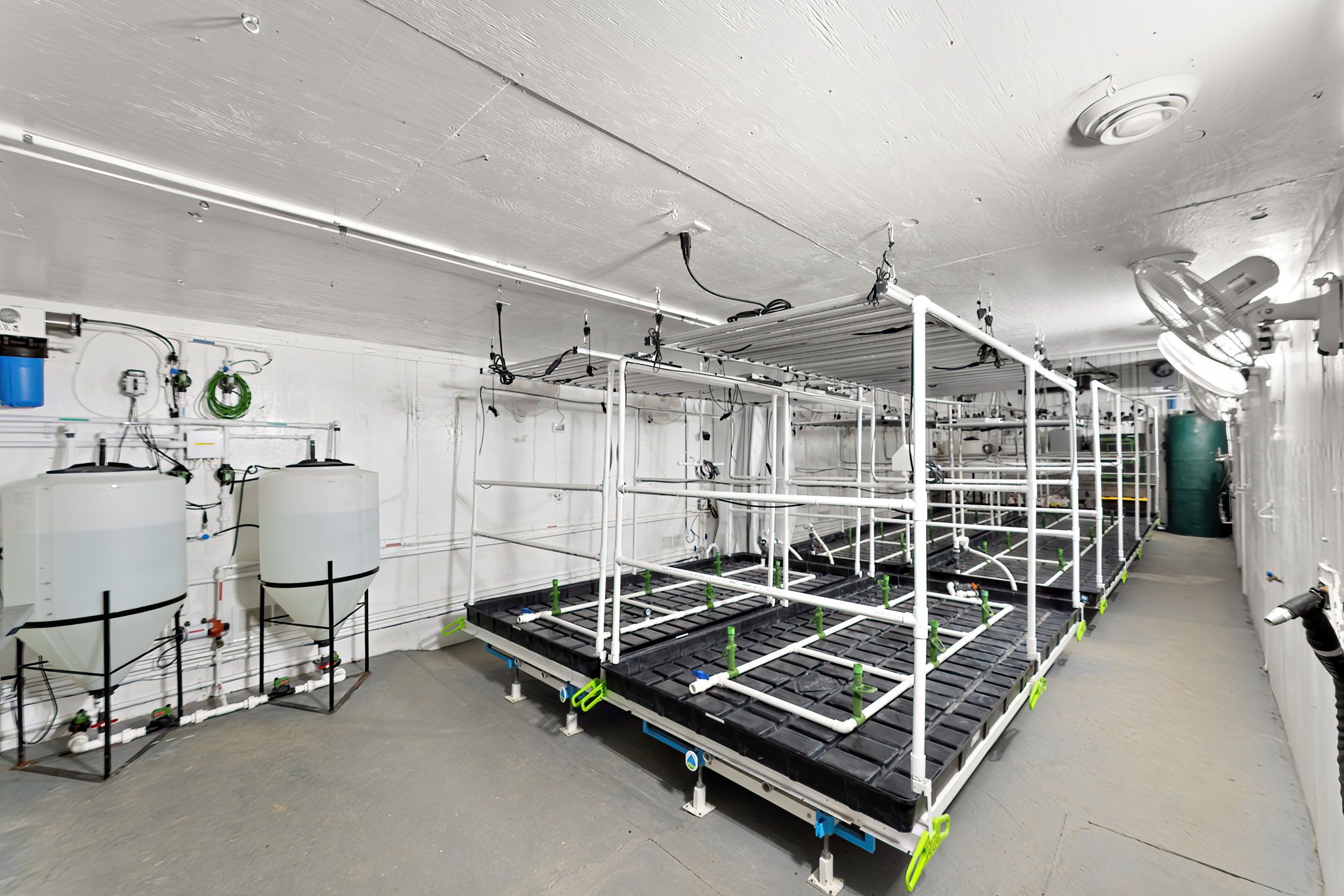 White-walled indoor hydroponic setup with multiple black growing trays, white PVC pipe frames, cables, large tanks on stands, and wall-mounted fans along a narrow corridor.