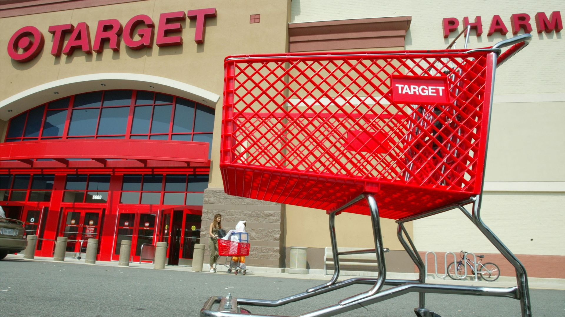 Close-up of an empty red Target shopping cart outside a Target store with red entrance doors and signage, a person pushing another cart is visible in the background.
