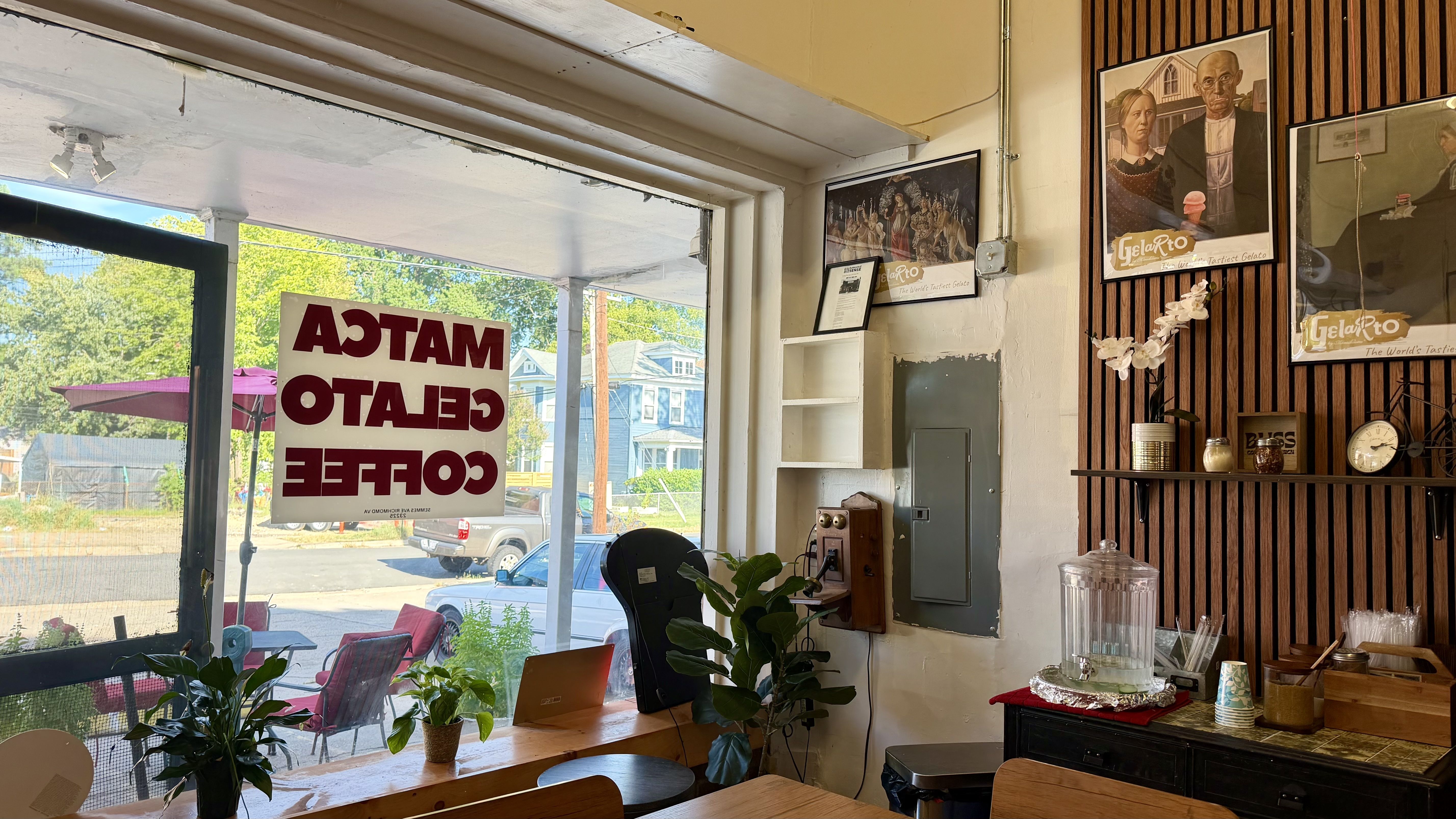 Cozy cafe interior with a wood wall and vintage posters, including Gelarto ads. Plants on the counter, a glass beverage dispenser, clock and jars. Window shows outdoor seating and reversed sign.