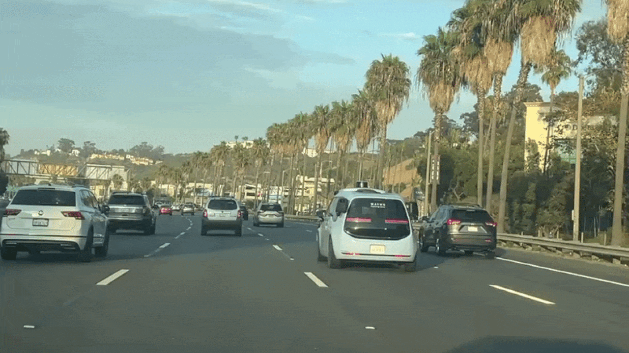 Sunny highway scene with multiple cars, including a white van in the foreground, driving along a road lined by tall palm trees on the right under a blue sky with light clouds.