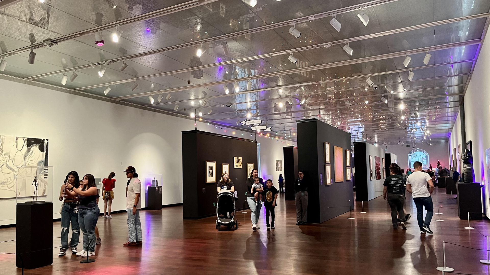 People stroll the gallery room, with features fall black walls of artwork and colorful string lights reflected on the ceiling.