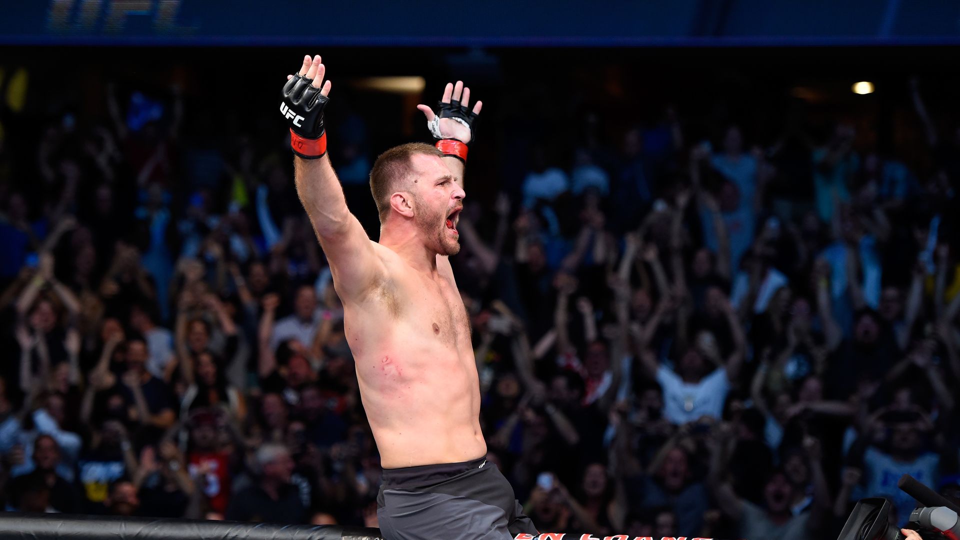 UFC fighter Stipe Miocic sits on top of the Octagon with his hands up in victory. 