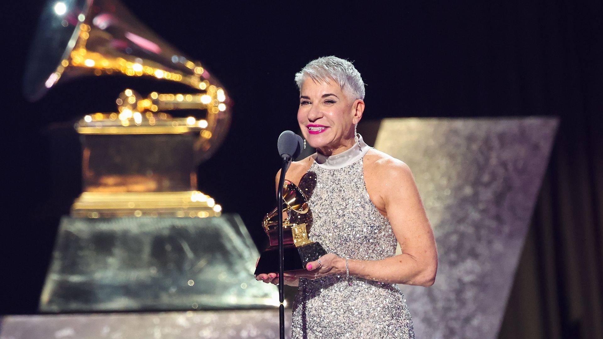 Woman with short silver hair in a sparkling silver dress holding a Grammy Award trophy and speaking at a microphone on stage with a large golden Grammy statue in the background.