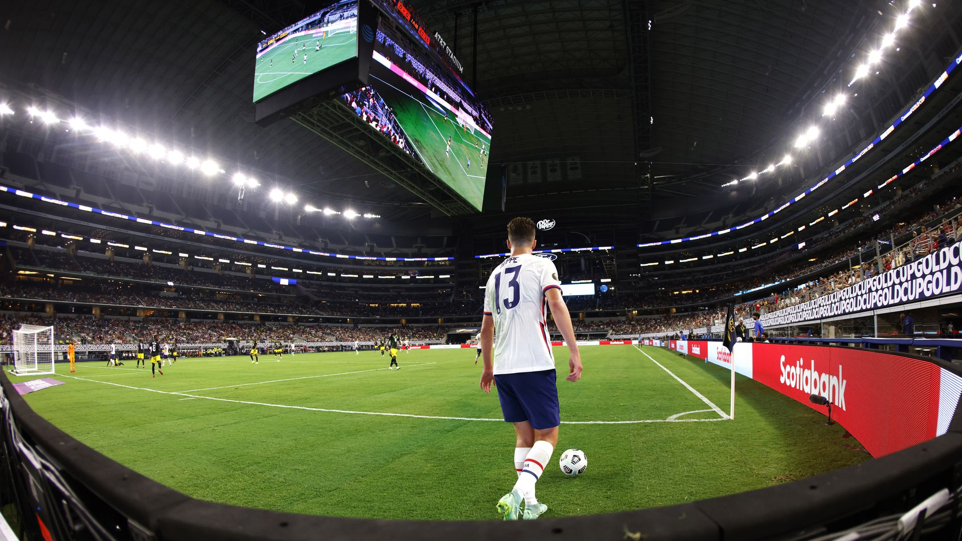 An American soccer player, wearing a white jersey, lines up for a corner kick under a large jumbotron.
