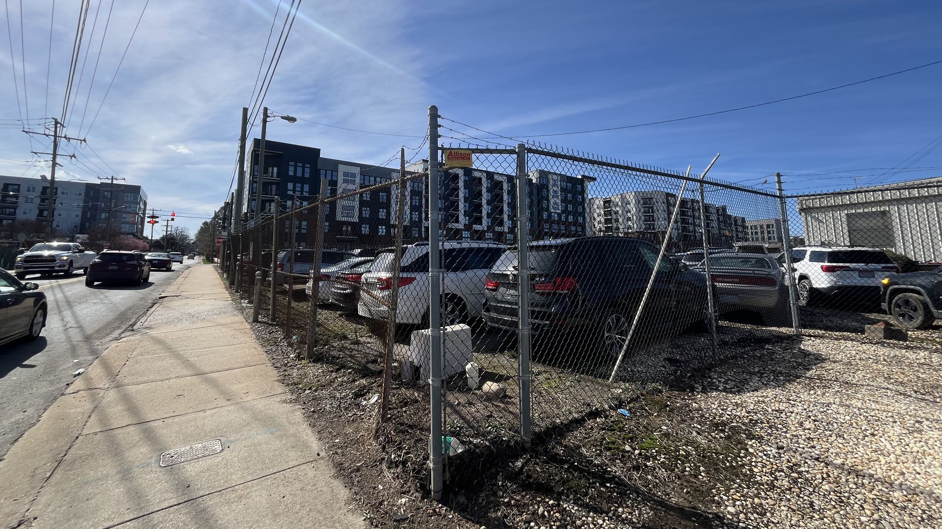 Daytime urban scene with a sidewalk next to a chain-link fence enclosing a gravel parking lot filled with cars. Modern multi-story buildings and blue sky with light clouds in the background.