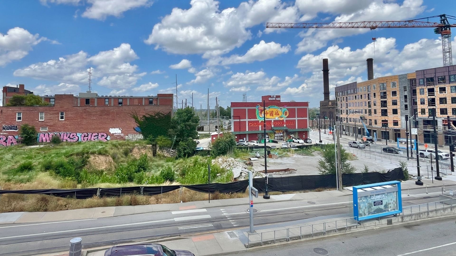 Left side of the photo shows an excavated trench in the ground with weeds and trees growing. To the right is a Chinese market and a crumbling parking lot, and on the right side of the picture is an apartment project in the middle of construction. The Streetcar stop on 3rd Street is in the foreground