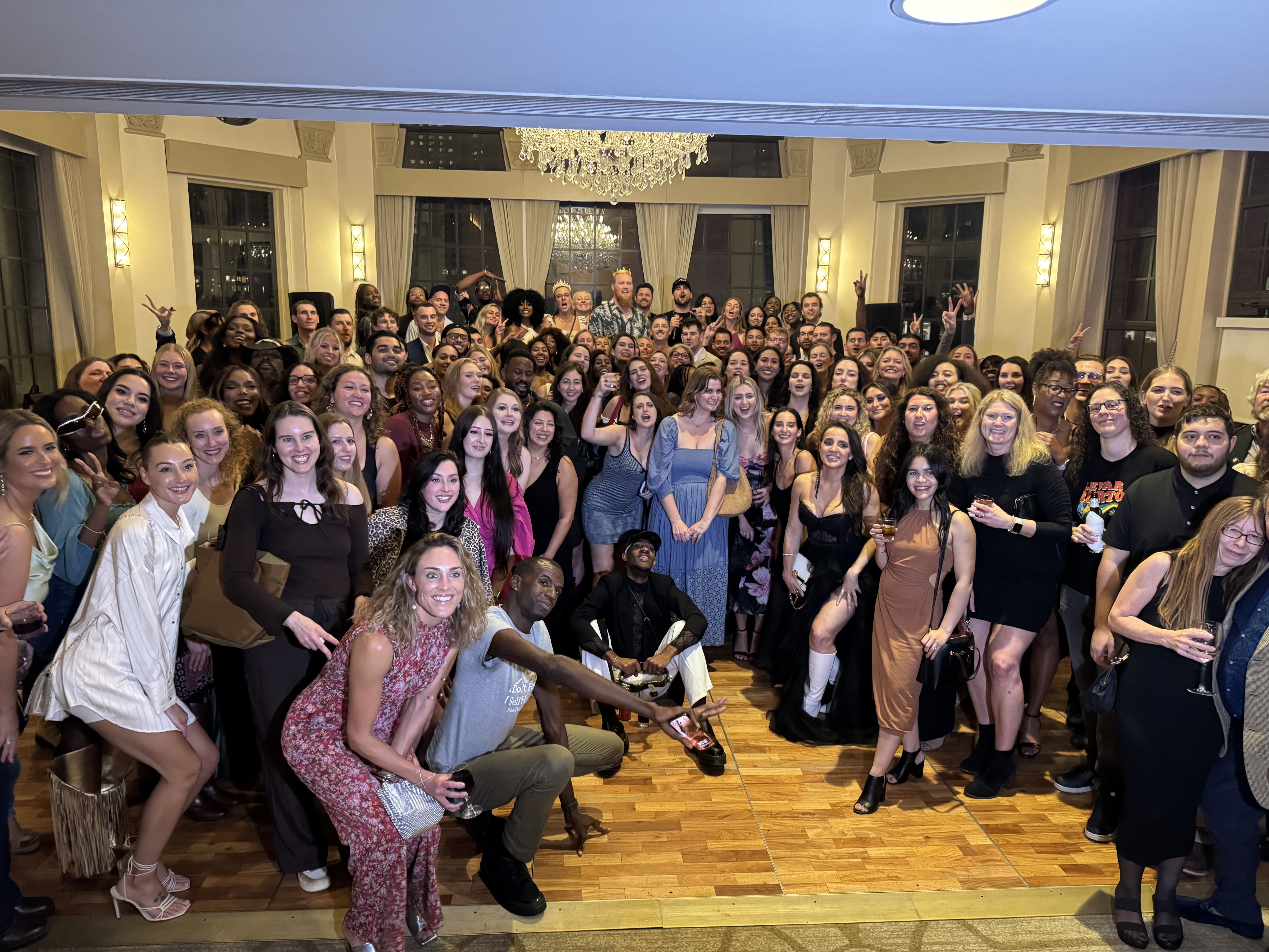 Large diverse group of people gathered indoors for a celebration, dressed in semi-formal to casual attire, smiling and posing under a chandelier in a room with large windows.