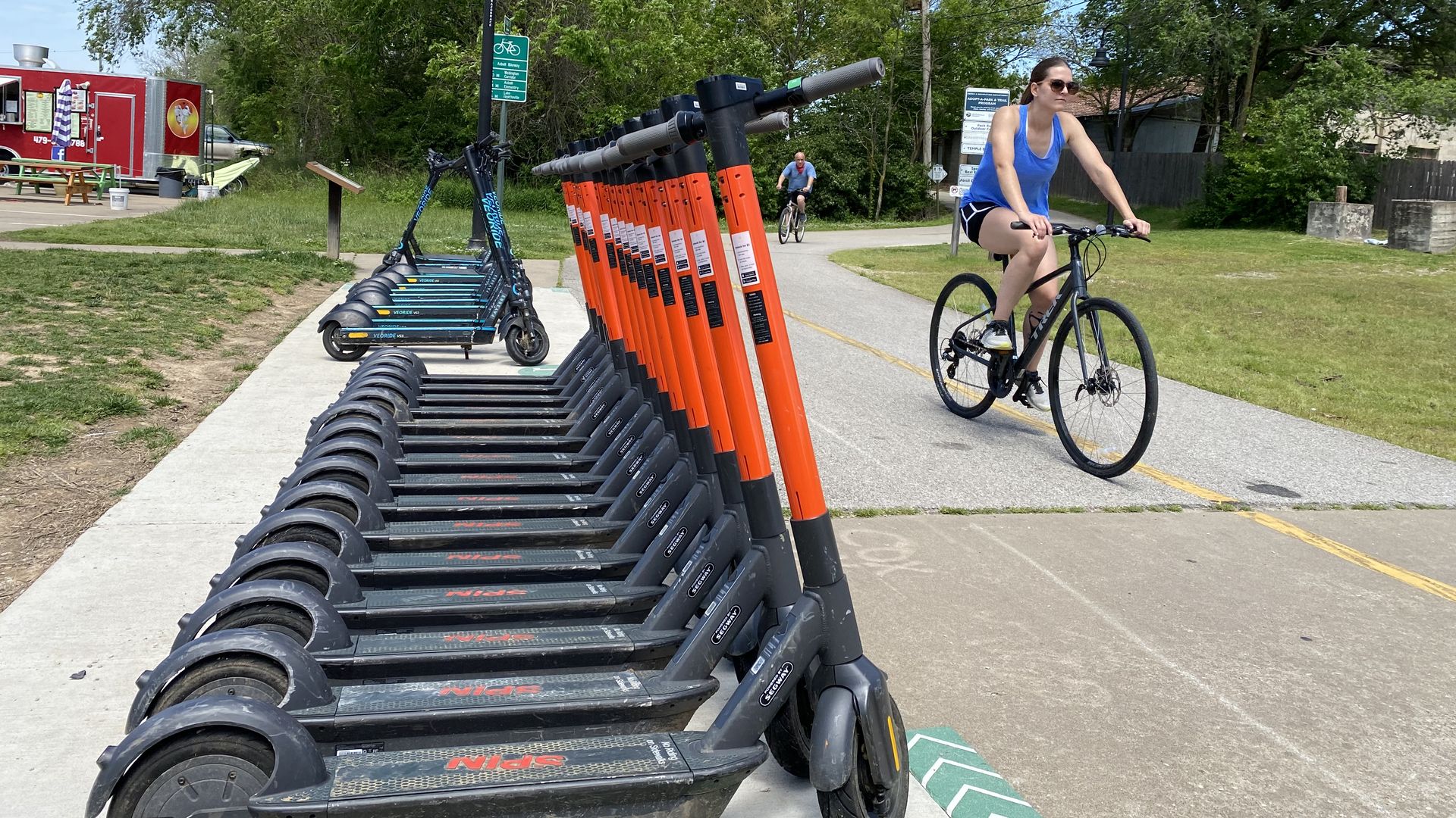 A cyclists rides past several e-scooters parked along the a trail. 