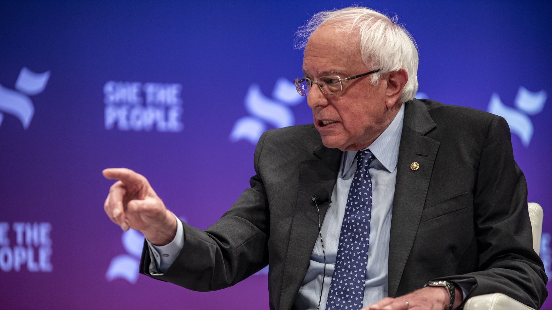 In this image, Bernie Sanders sits in a chair and talks while pointing a finger at something.