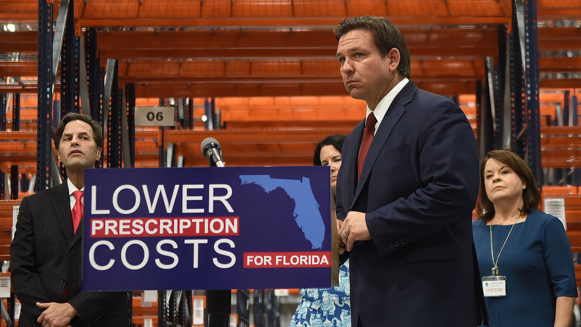Picture of Ron DeSantis standing next to a sign that says "lower prescription drugs"