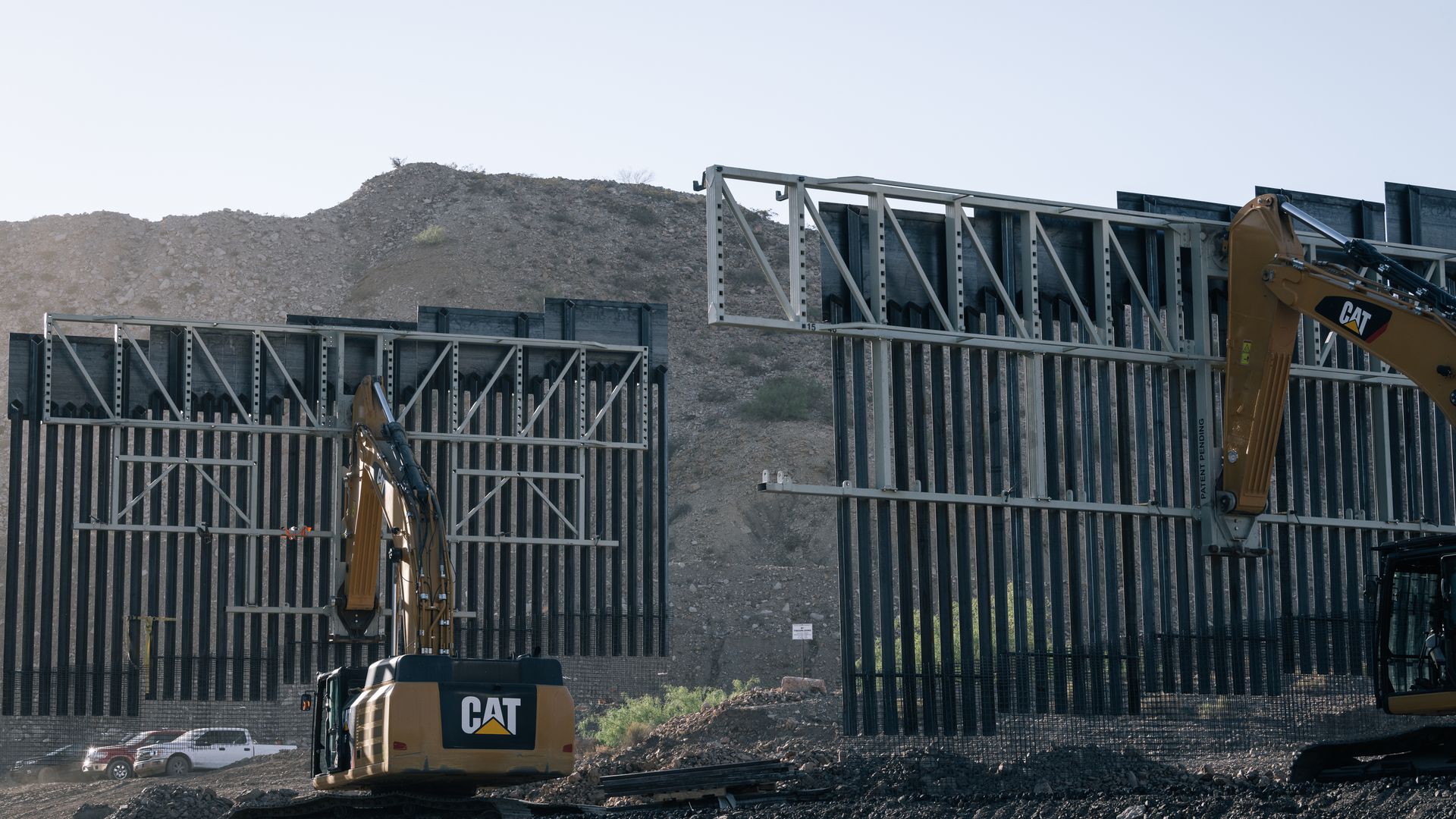 Construction equipment lowers big portions of slatted fence border wall into the ground. Mountains are in the background.