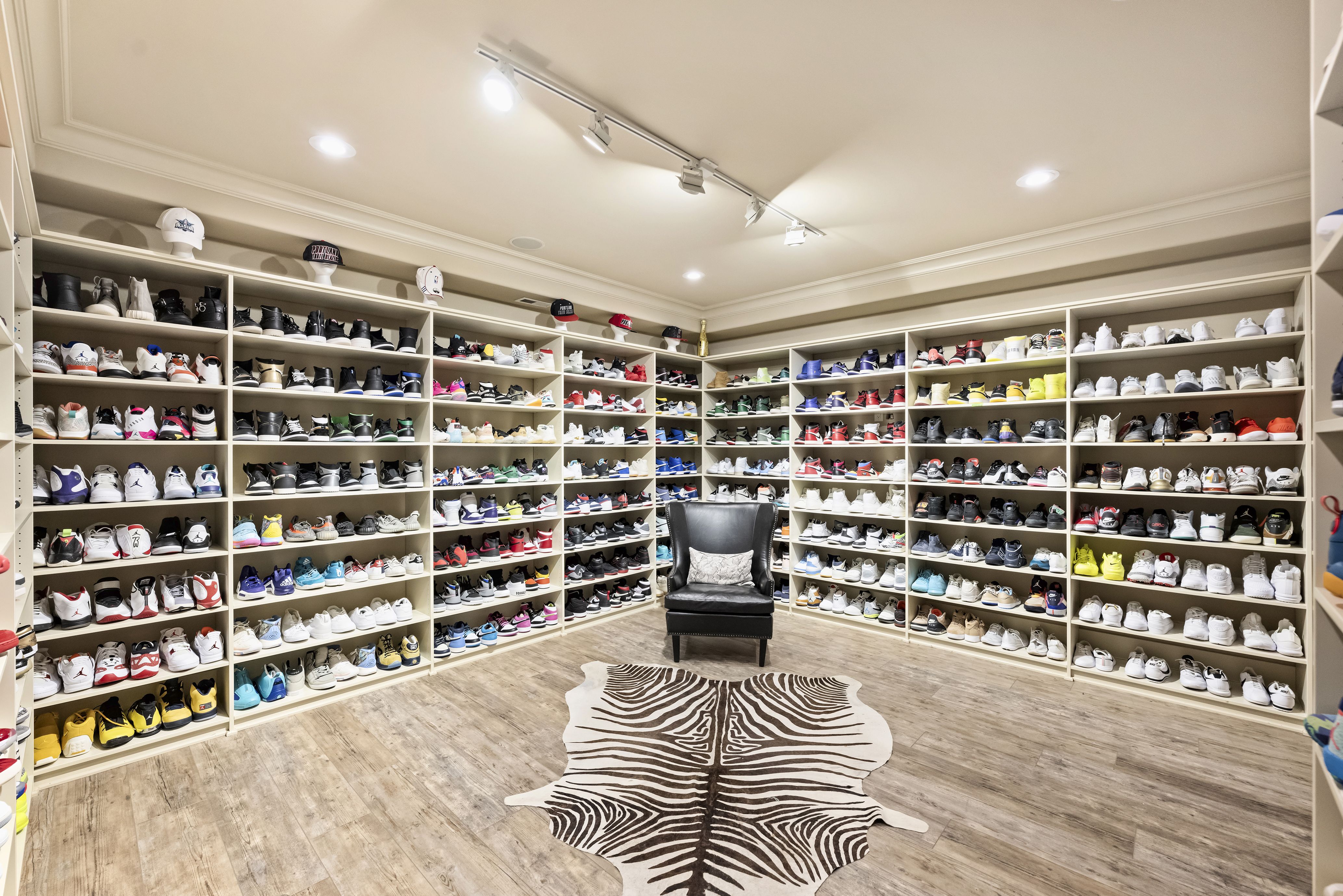 Spacious walk-in shoe closet with white shelves full of sneakers, white hats on top, a black chair in the center, zebra rug on wooden floor, and track lighting overhead.