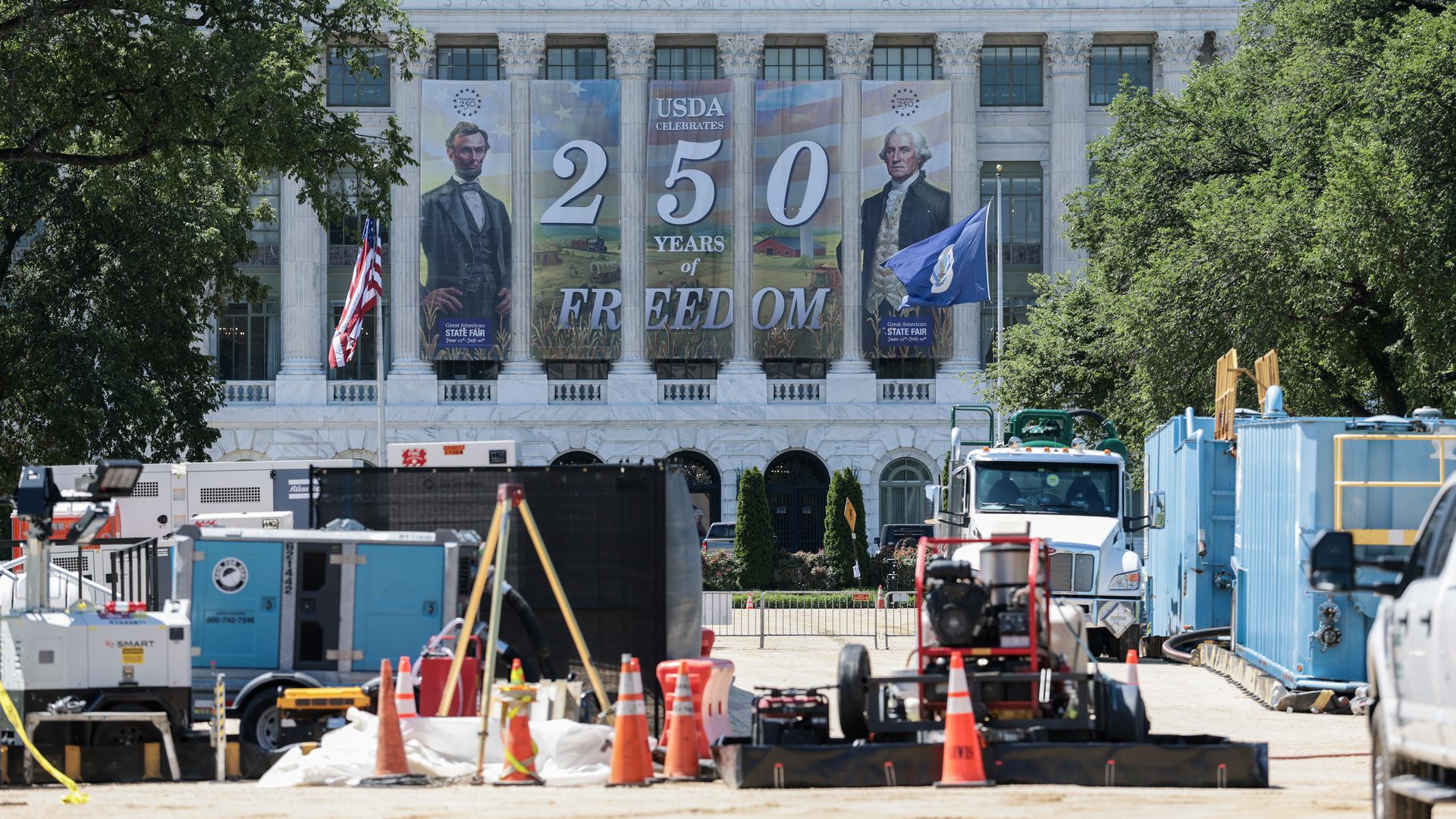 View of a marble government building with large banners reading 250 Years of Freedom and portraits, flanked by trees; foreground shows a busy construction site with orange cones and equipment.