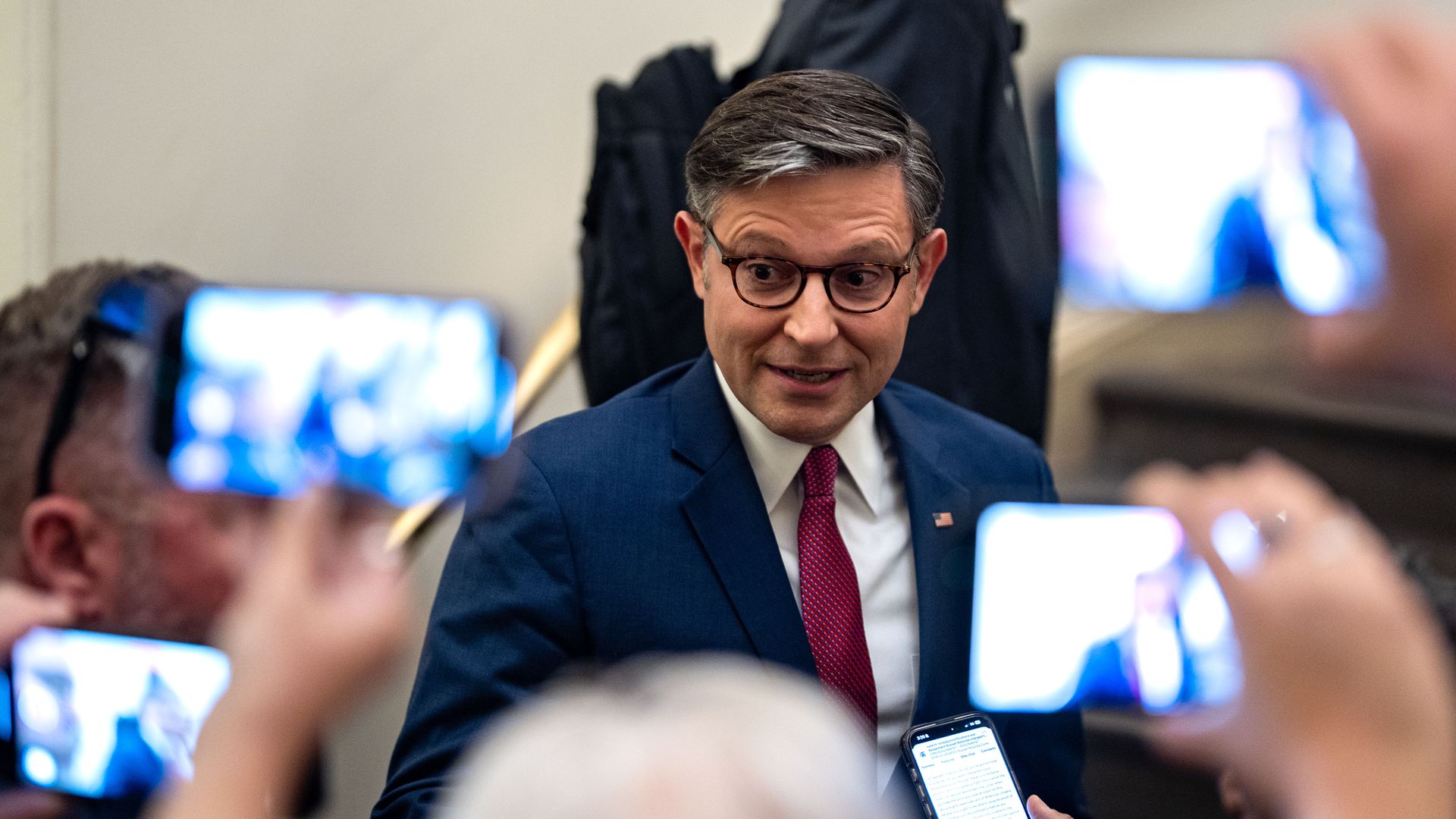 Reporters hold up their phones to film Mike Johnson, wearing a blue suit in front of a white wall.