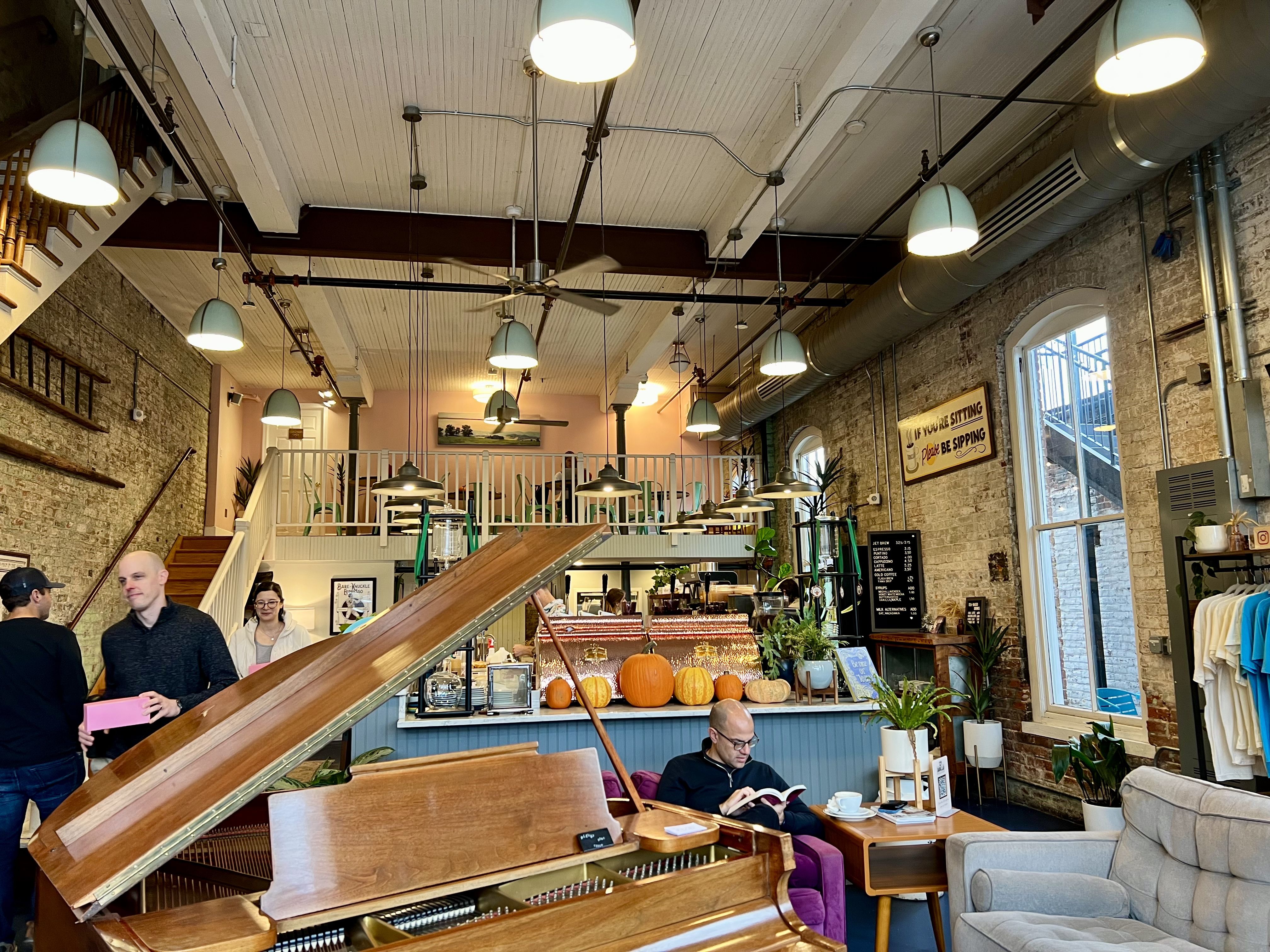 The inside of a coffee shop with two levels. The first floor has a brown wooden piano and someone reading beside it in a purple chair. There are pumpkins lining the serving area. The second floor has a bunch of chairs.